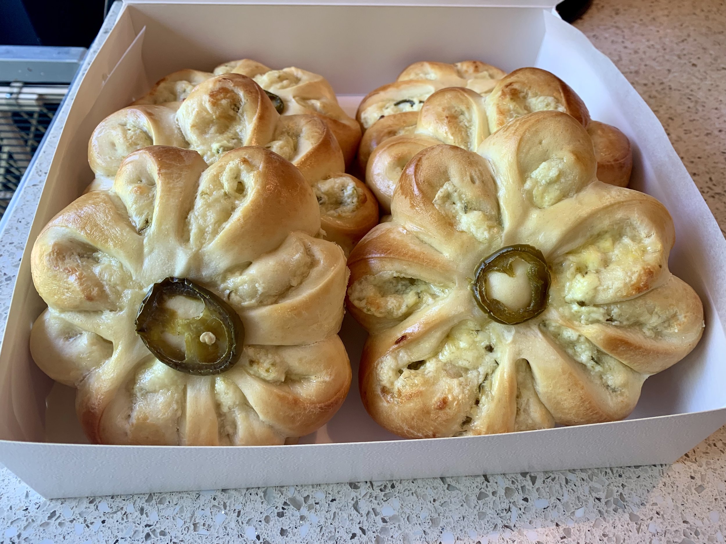 A white box containing two baked bread rolls shaped like flowers, each topped with a slice of jalapeƱo pepper.