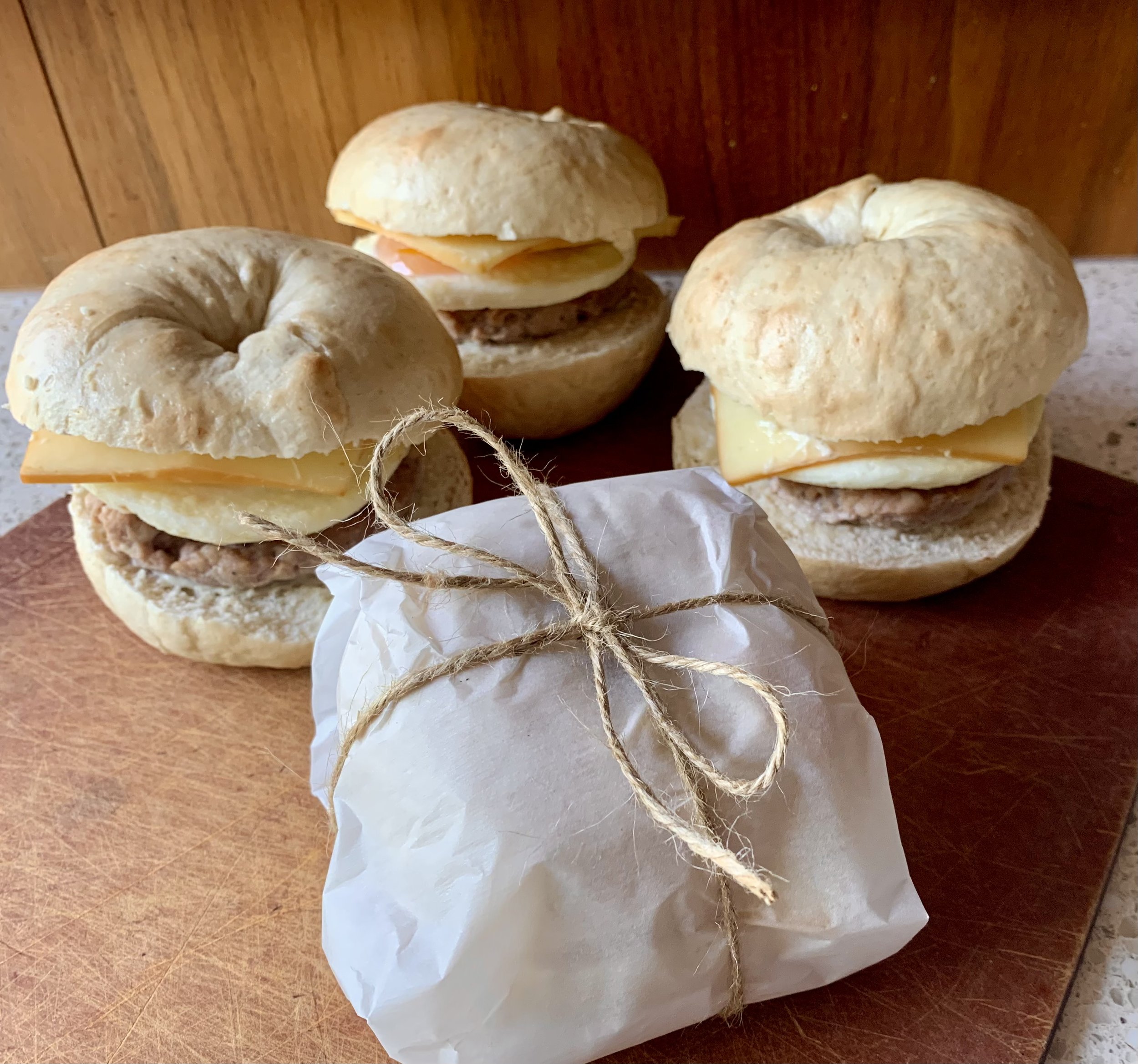 Three breakfast sandwiches made with bagels, cheese, sausage, and eggs on a wooden cutting board. There is also a wrapped sandwich tied with string.