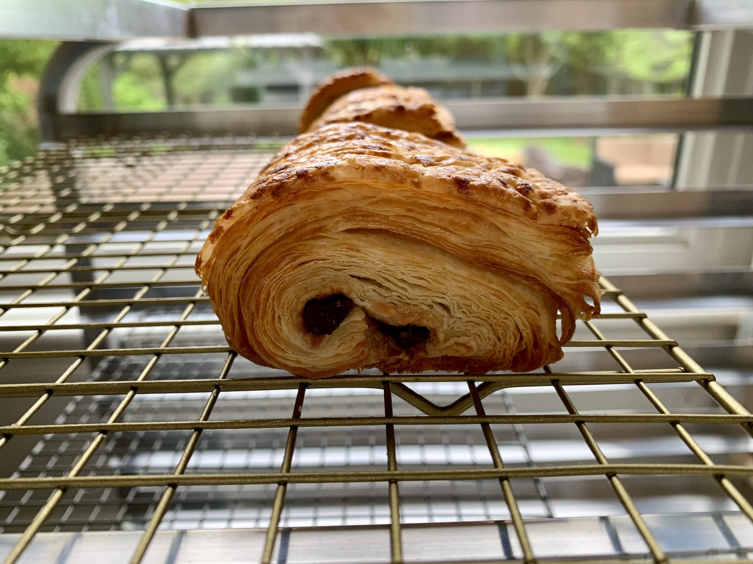 A freshly baked pain au chocolat pastry with visible chocolate inside, resting on a metal cooling rack near a window with green outdoor scenery.