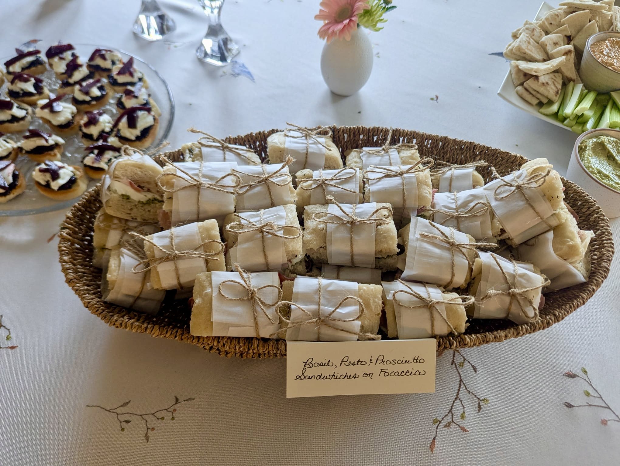 A wicker basket filled with small sandwiches wrapped in white paper and tied with twine, labeled 'Basil Pesto and Prosciutto Sandwiches on Focaccia'. Surrounding the basket are plates of mini desserts, and a bowl of dip.