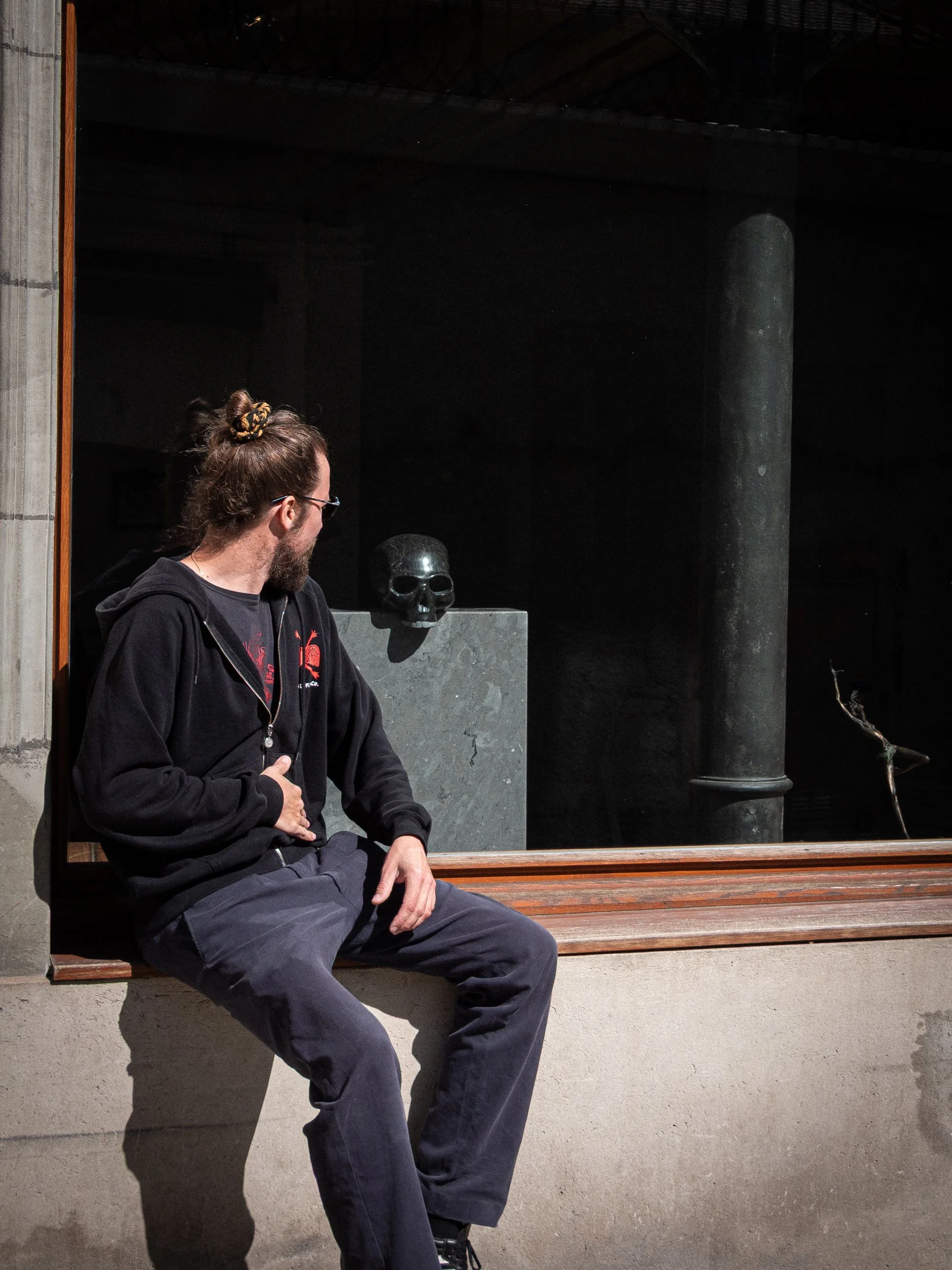 Homme avec des cheveux attachés et une barbe assis en regardant par une fenêtre, à côté d'une sculpture de crâne en noir dans un environnement sombre