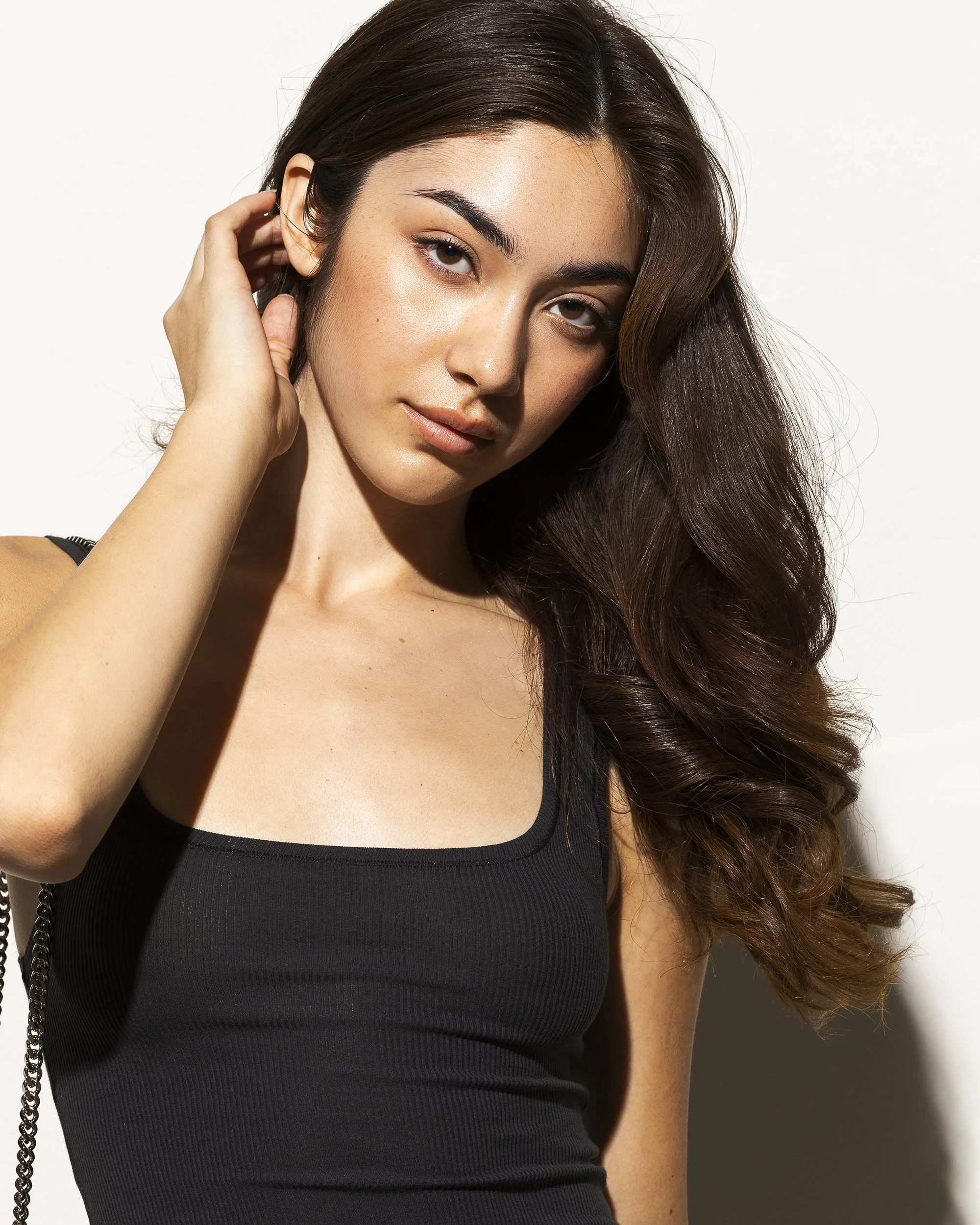 A young woman with long, wavy brown hair, wearing a black tank top, posing in front of a plain white background.