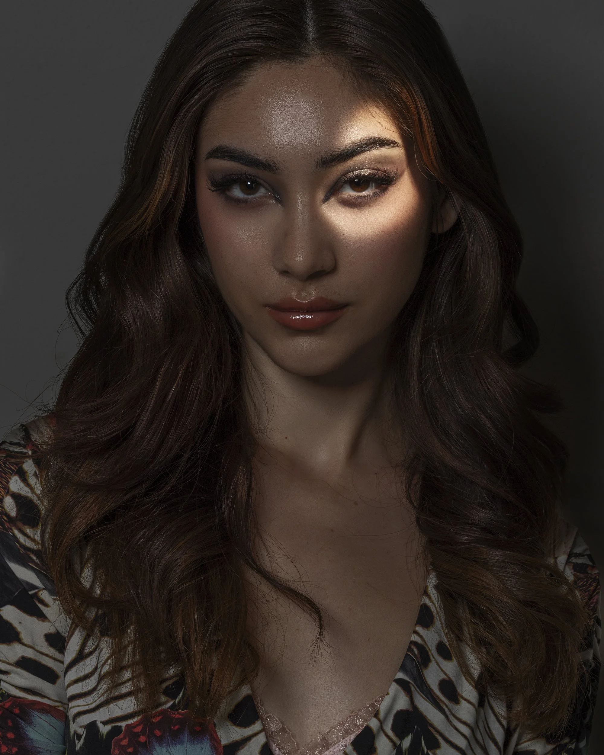 A woman with long, wavy brown hair, wearing makeup, a slight smile, wearing a colorful patterned top, illuminated with dramatic lighting.