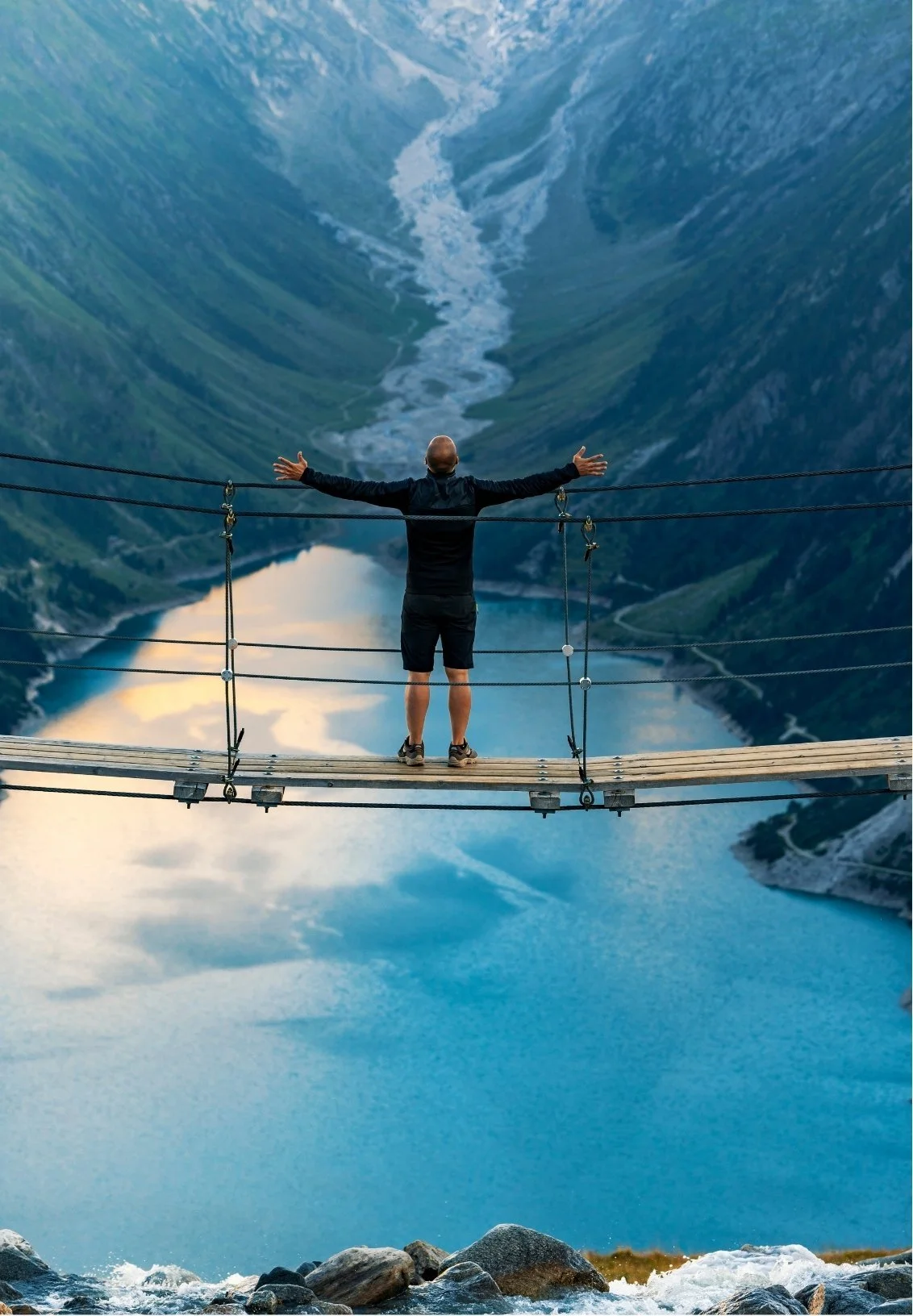 A man standing on a suspension bridge with arms stretched wide, overlooking a deep valley with a turquoise lake and steep mountain slopes.