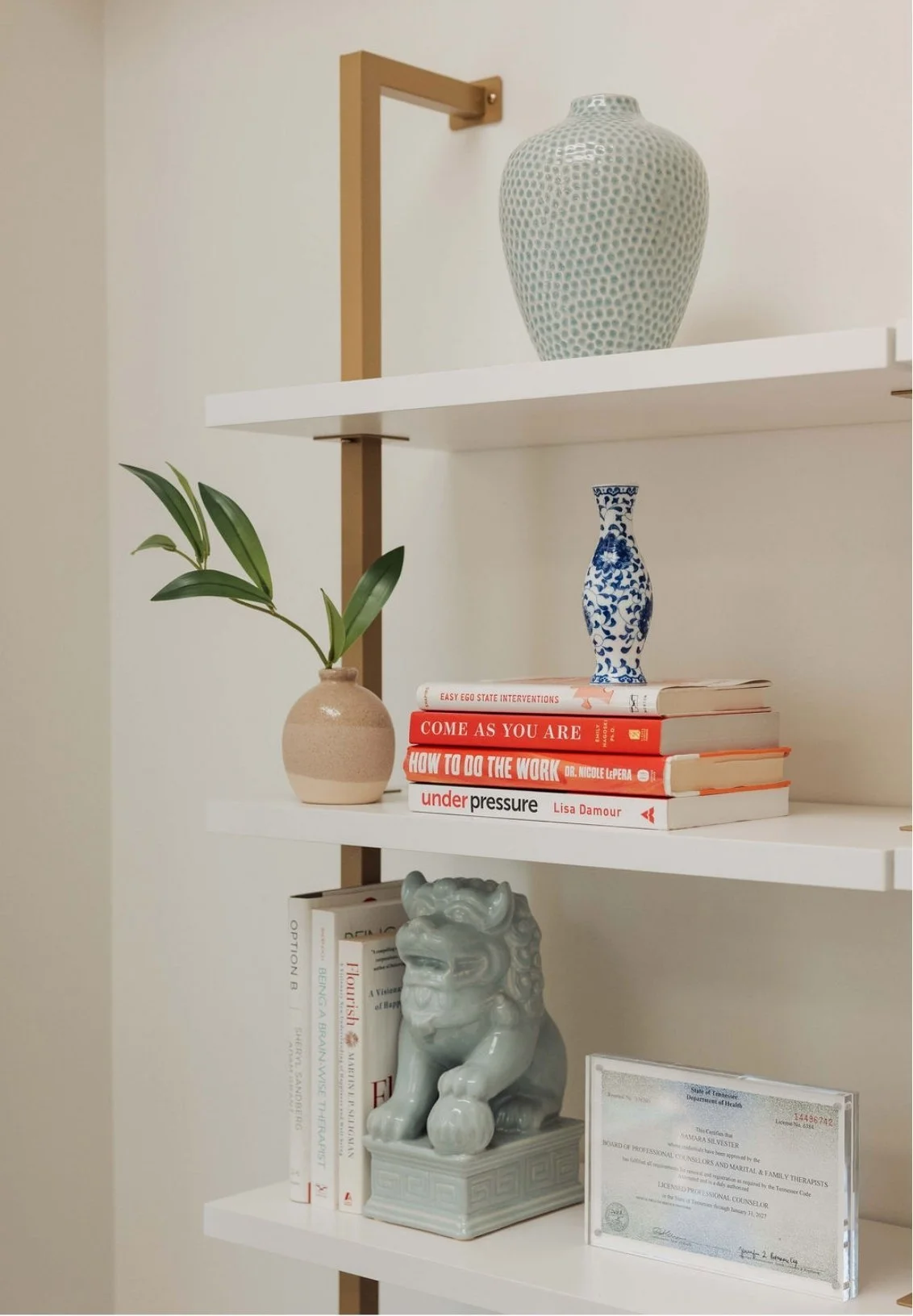 White bookshelf in a Nashville therapy office with decorative vases, books, a ceramic lion, and a framed professional license, against a light-colored wall.