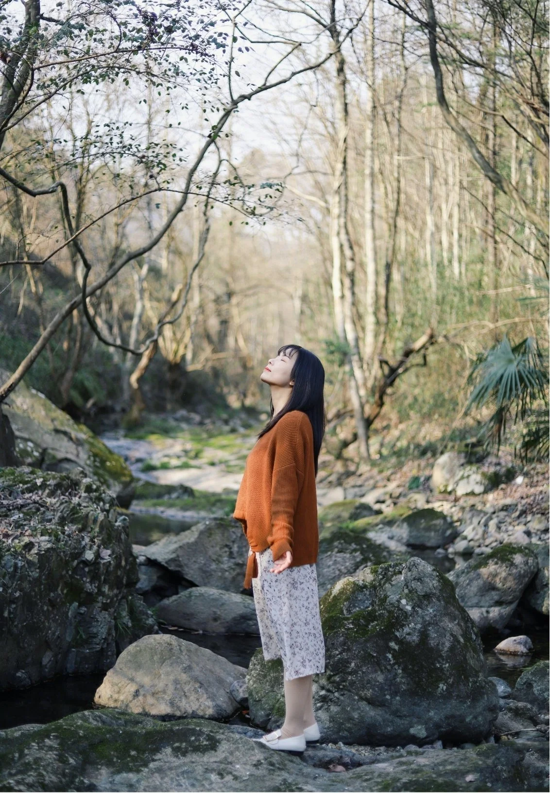 A woman standing on rocks in a forested area with a stream, her eyes closed and face tilted upward, wearing a brown sweater, patterned skirt, and white shoes.