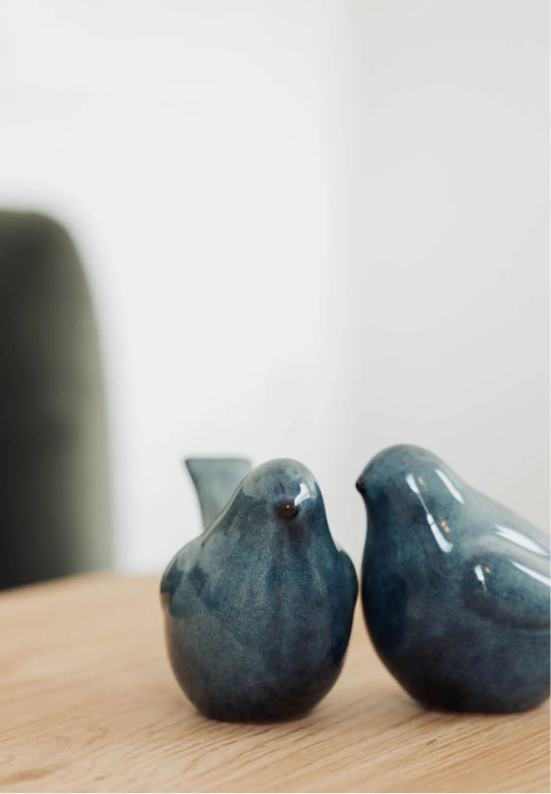 Two ceramic bird figurines, one smaller and one larger, with a blue glaze, sitting on a wooden surface. In the background, there is a blurred dark-colored object.