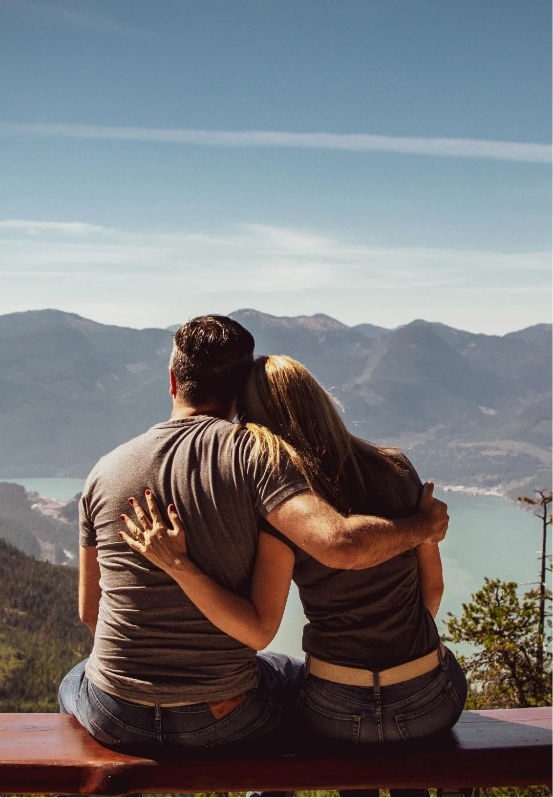 A couple sitting on a wooden bench, embracing and looking at a scenic mountain landscape with a lake in the distance.