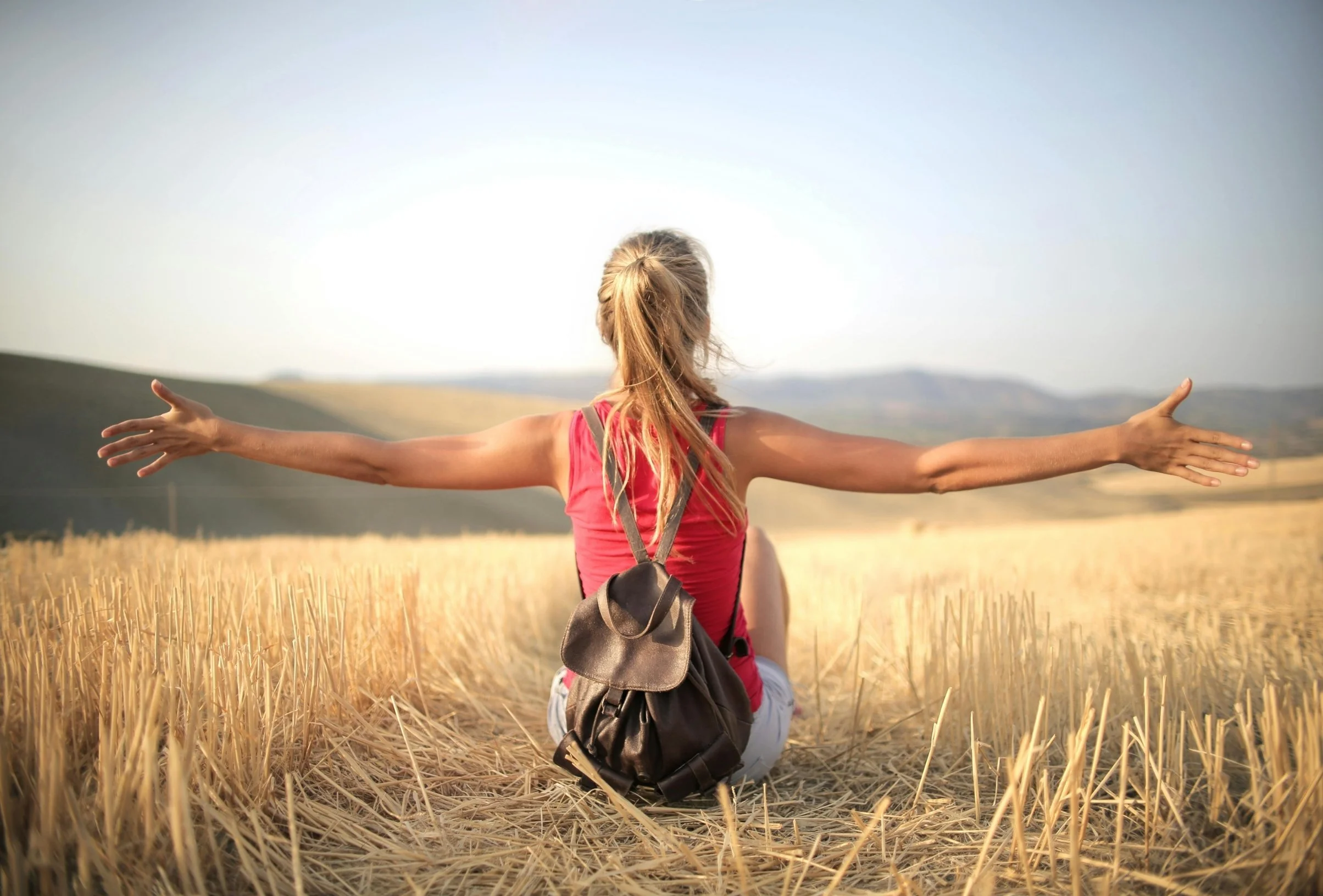 Woman sitting in a wheat field with arms outstretched, backpack on her back, facing away, enjoying a rural landscape.