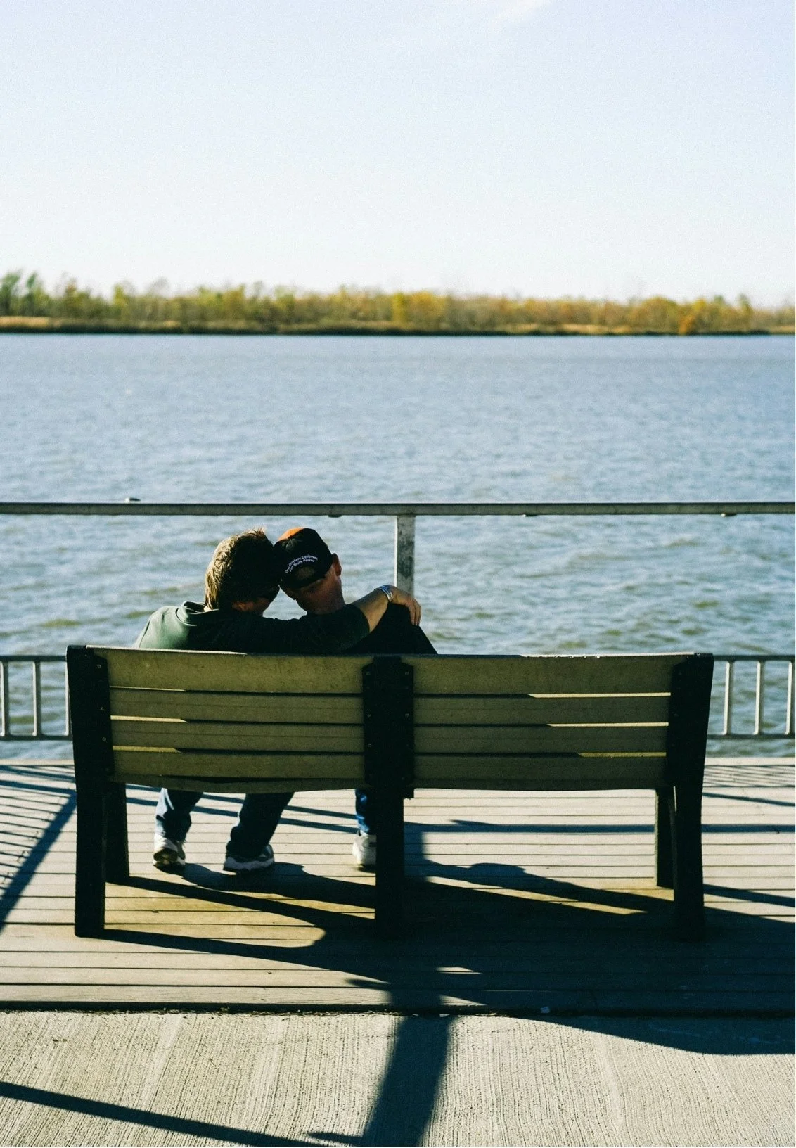 Two people sitting closely on a bench by the water, with one person resting their head on the other's shoulder, enjoying a peaceful moment.