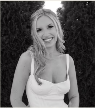 A woman with long wavy hair smiling at the camera, wearing a sleeveless top, outdoors with trees in the background.