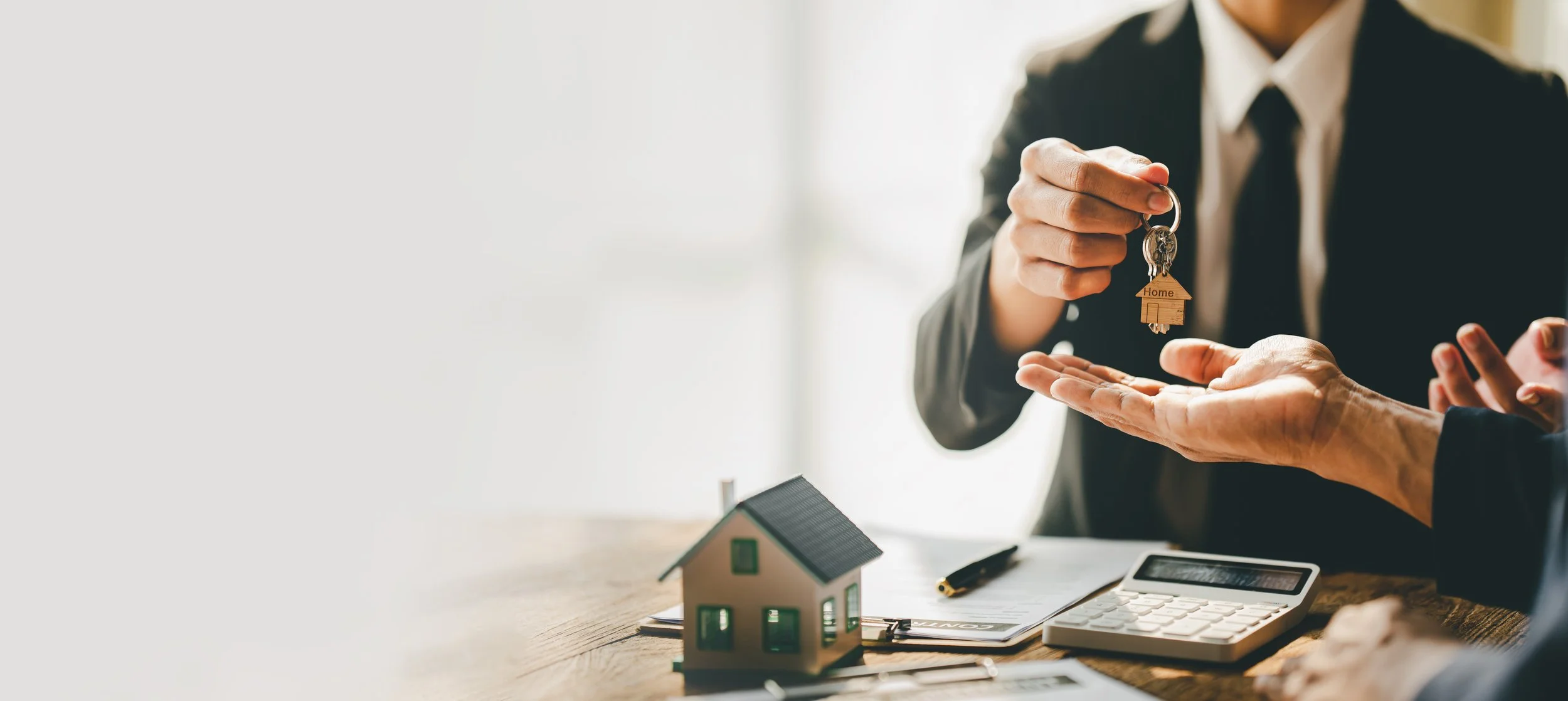 Person handing over house keys with a wooden house model and a small house on the table, during a real estate transaction.