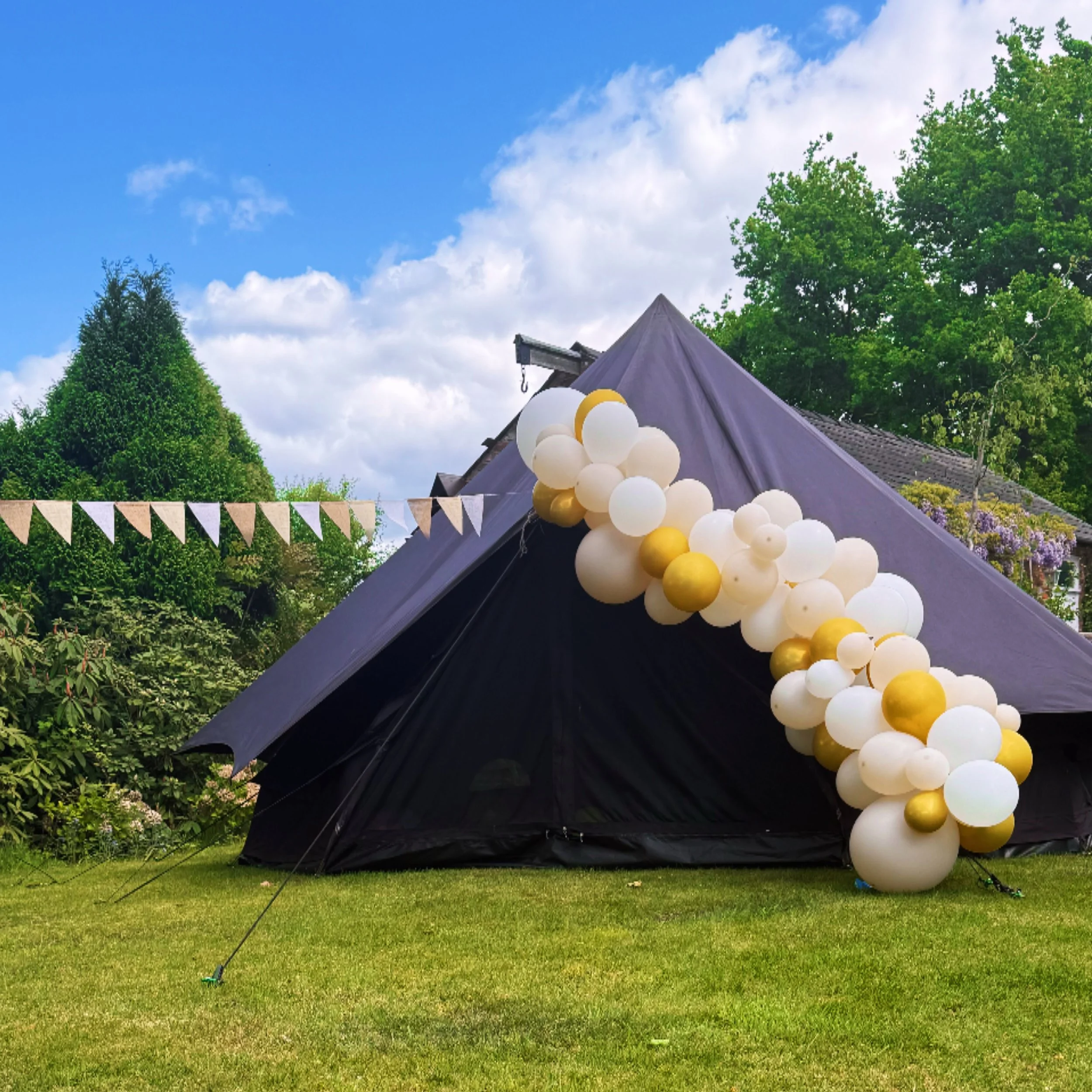 A black outdoor tent decorated with white, gold, and beige balloons, set up on a grassy area with trees and a house in the background under a partly cloudy sky.