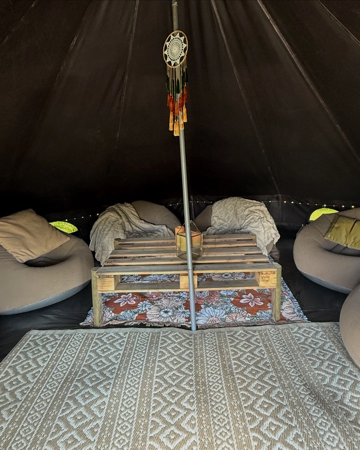 Inside a cozy tipi tent with a black fabric interior, featuring round bean bag chairs with cushions, a wooden pallet coffee table on a floral rug, a string of small lights along the tent wall, and a dream catcher hanging from a pole in the center.