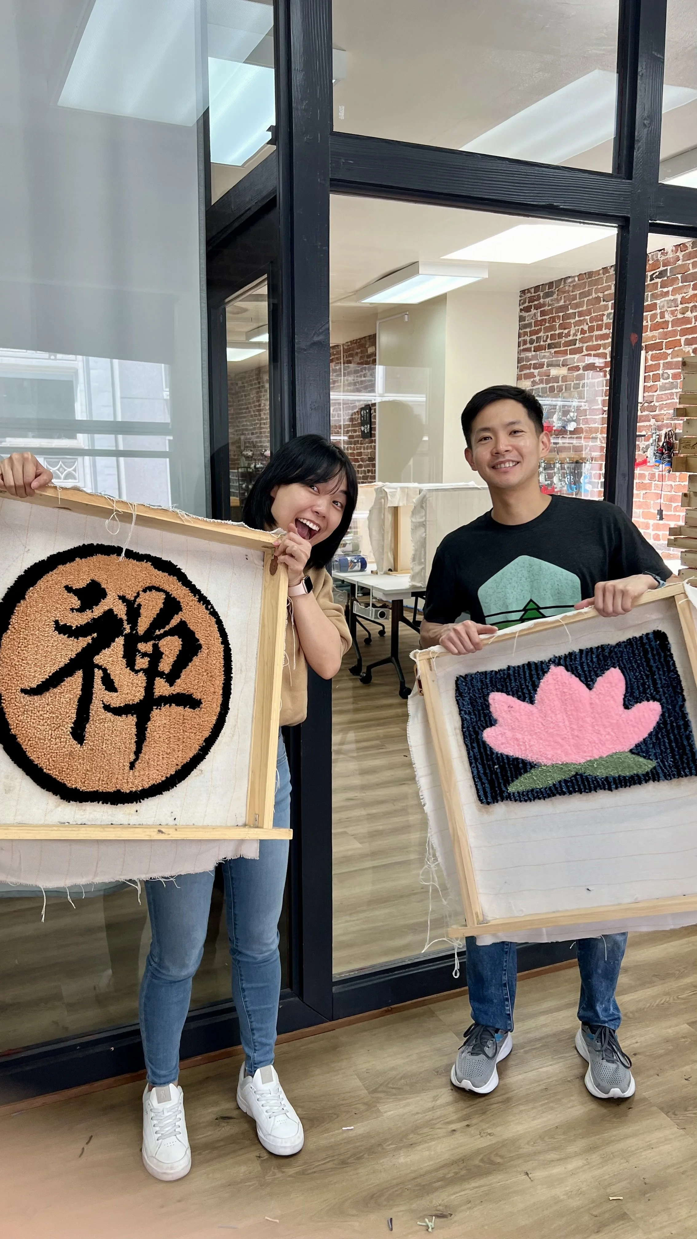 A woman and a man standing indoors, each holding a framed piece of textile art. The woman is smiling widely while the man is smiling calmly. The textile art includes a black and beige circle with a Chinese character and a pink flower on a black backg