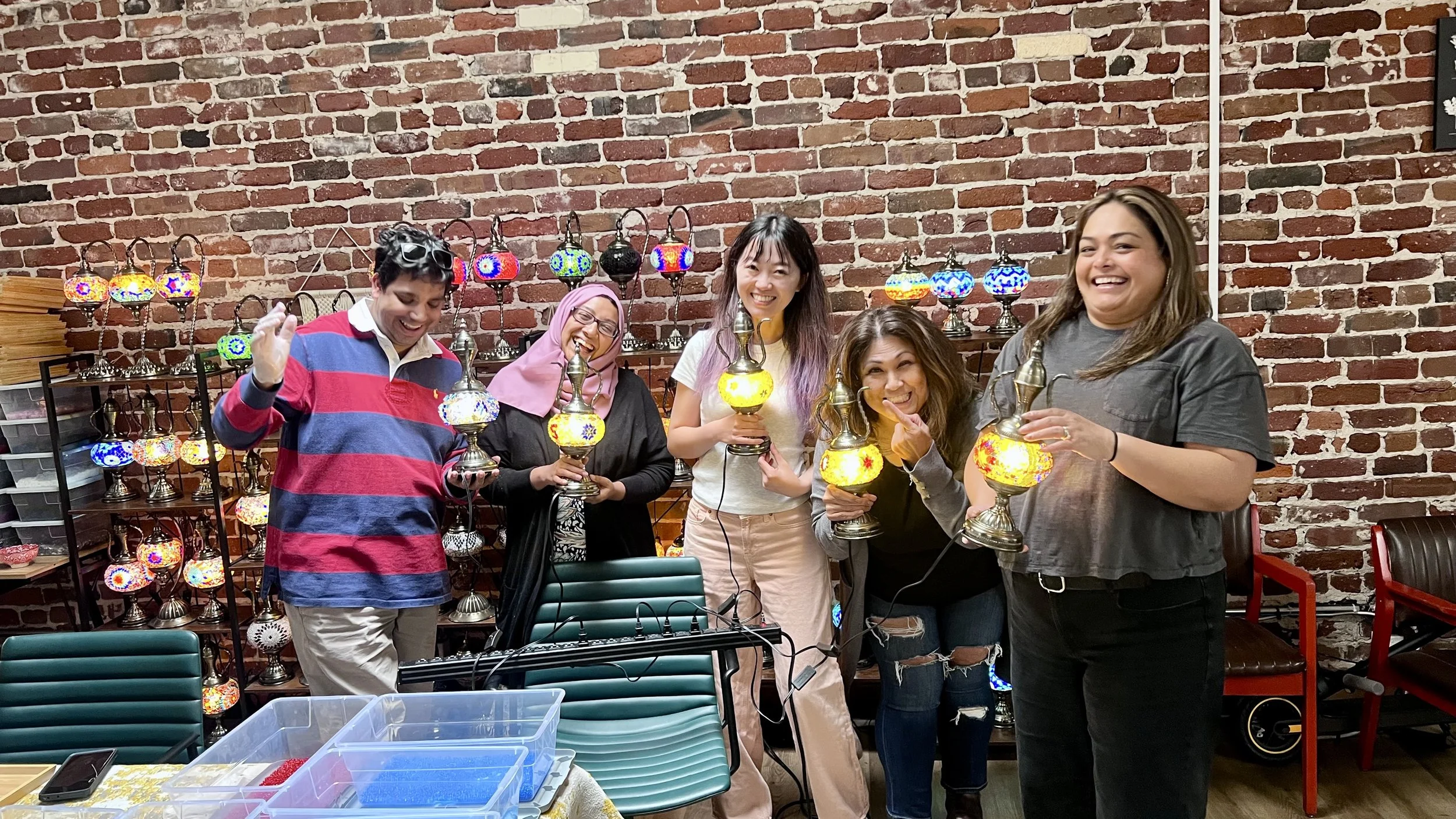 Group of five women smiling and holding colorful mosaic lamps in a workshop with a brick wall background and shelves of lamps behind them.