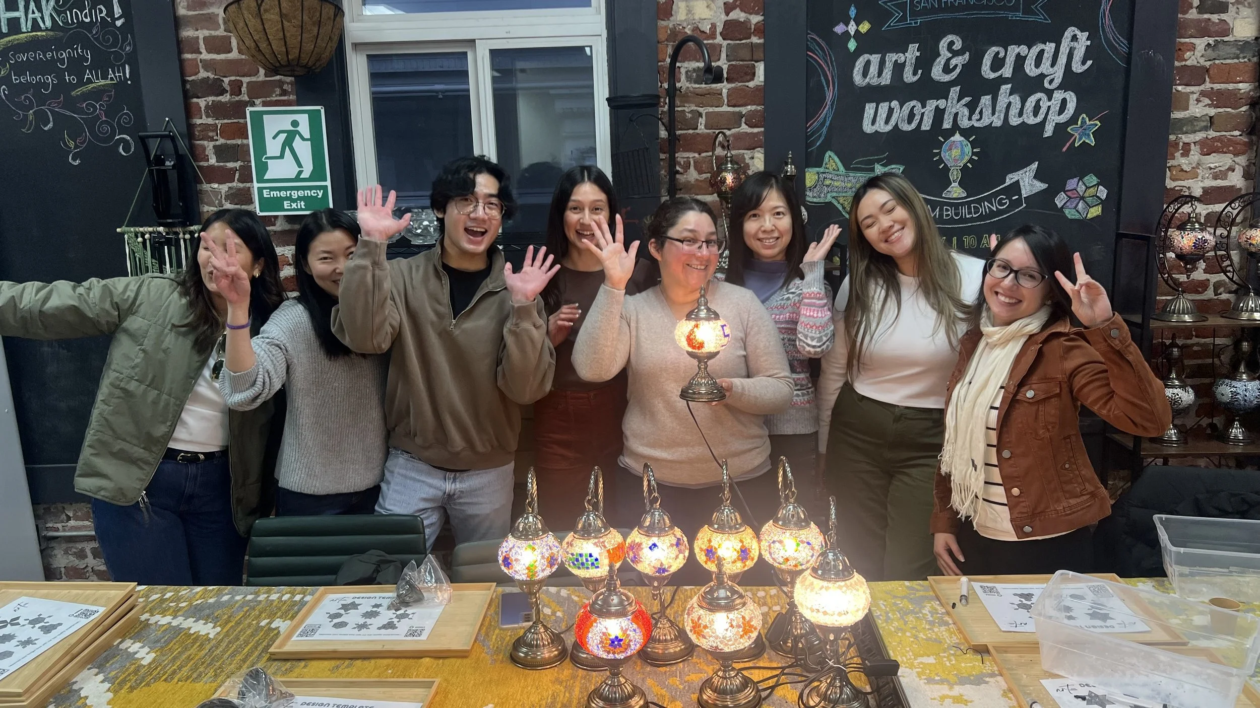 A group of eight people standing behind a table with colorful mosaic lamps, smiling and waving at the camera inside an art and craft workshop with brick walls and blackboards in the background.