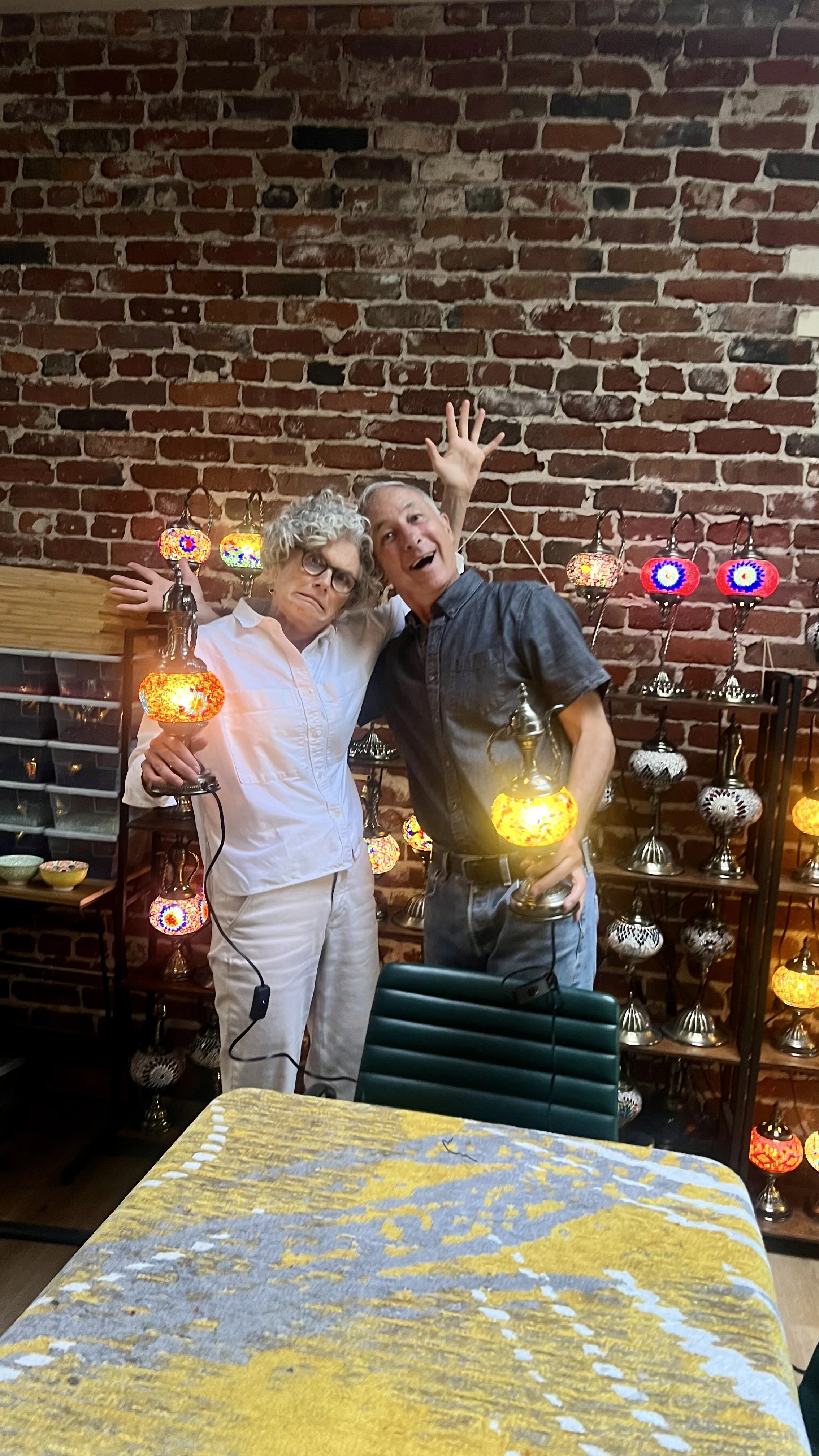 Two elderly people, a woman with curly gray hair and glasses, and a man with short gray hair, are posing playfully indoors in front of a display of colorful mosaic lamps on metal shelves against a brick wall. The woman looks amused while the man is s