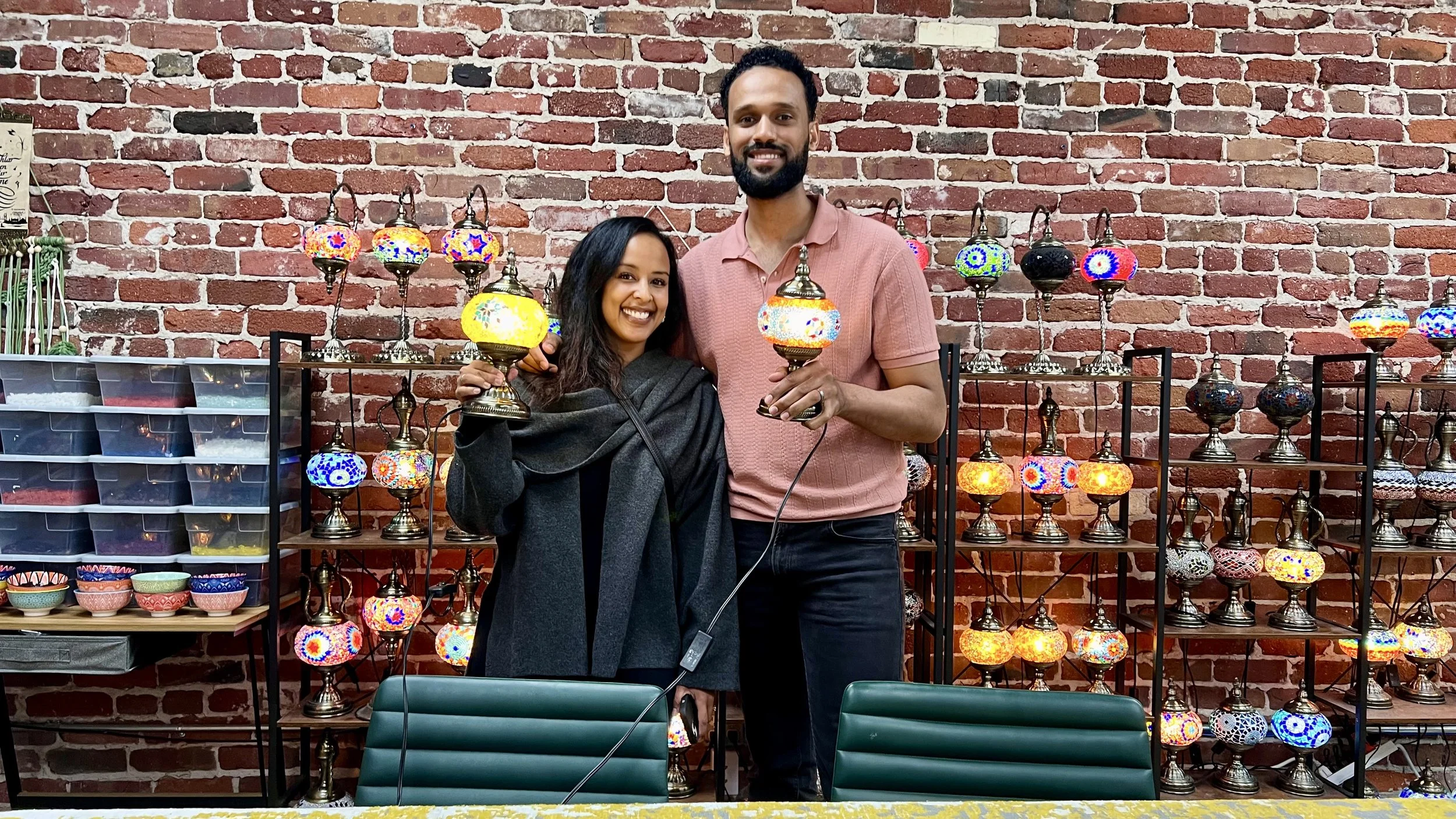 A man and woman smiling and holding colorful mosaic lamps in a store with shelves of mosaic lamps and pottery against a brick wall.