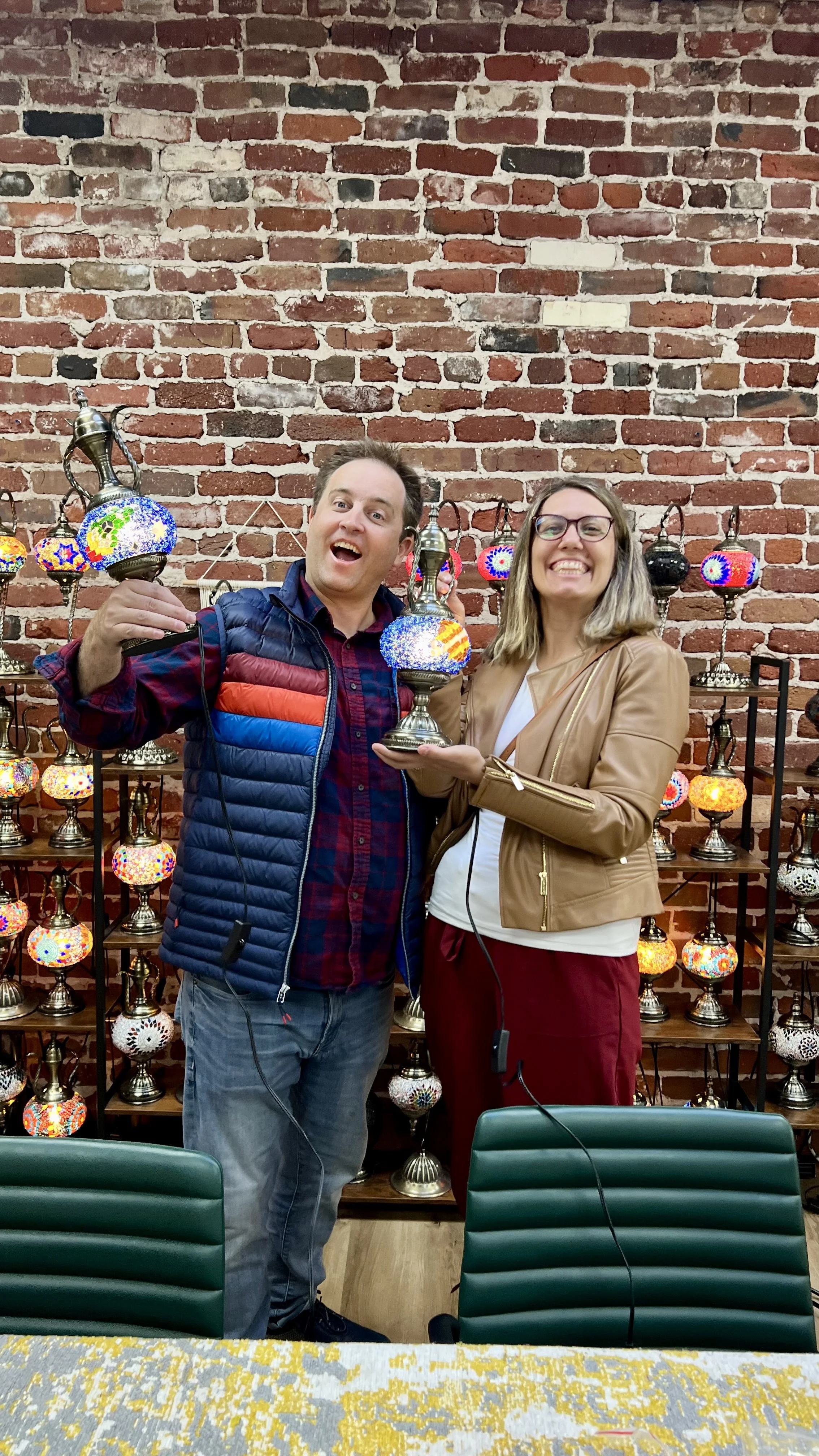 Two people smiling and holding Turkish mosaic lamps in front of a brick wall with shelves of colorful mosaic lamps.