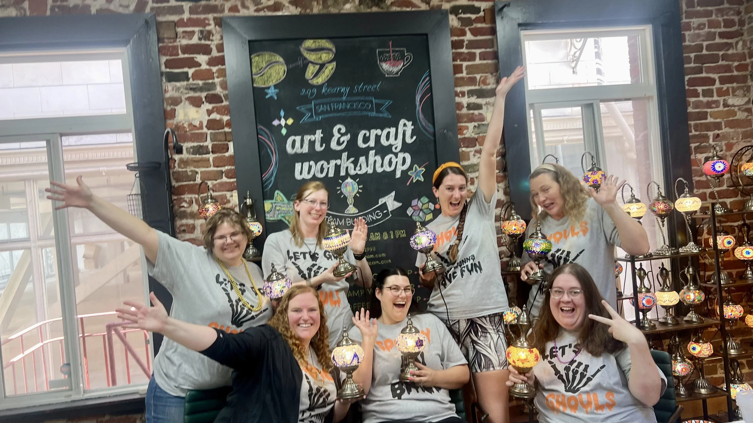 Group of seven women at an art and craft workshop, holding colorful mosaic lamps and smiling, inside a brick-walled studio with a chalkboard sign that reads 'art & craft workshop' and 'team building' in the background.