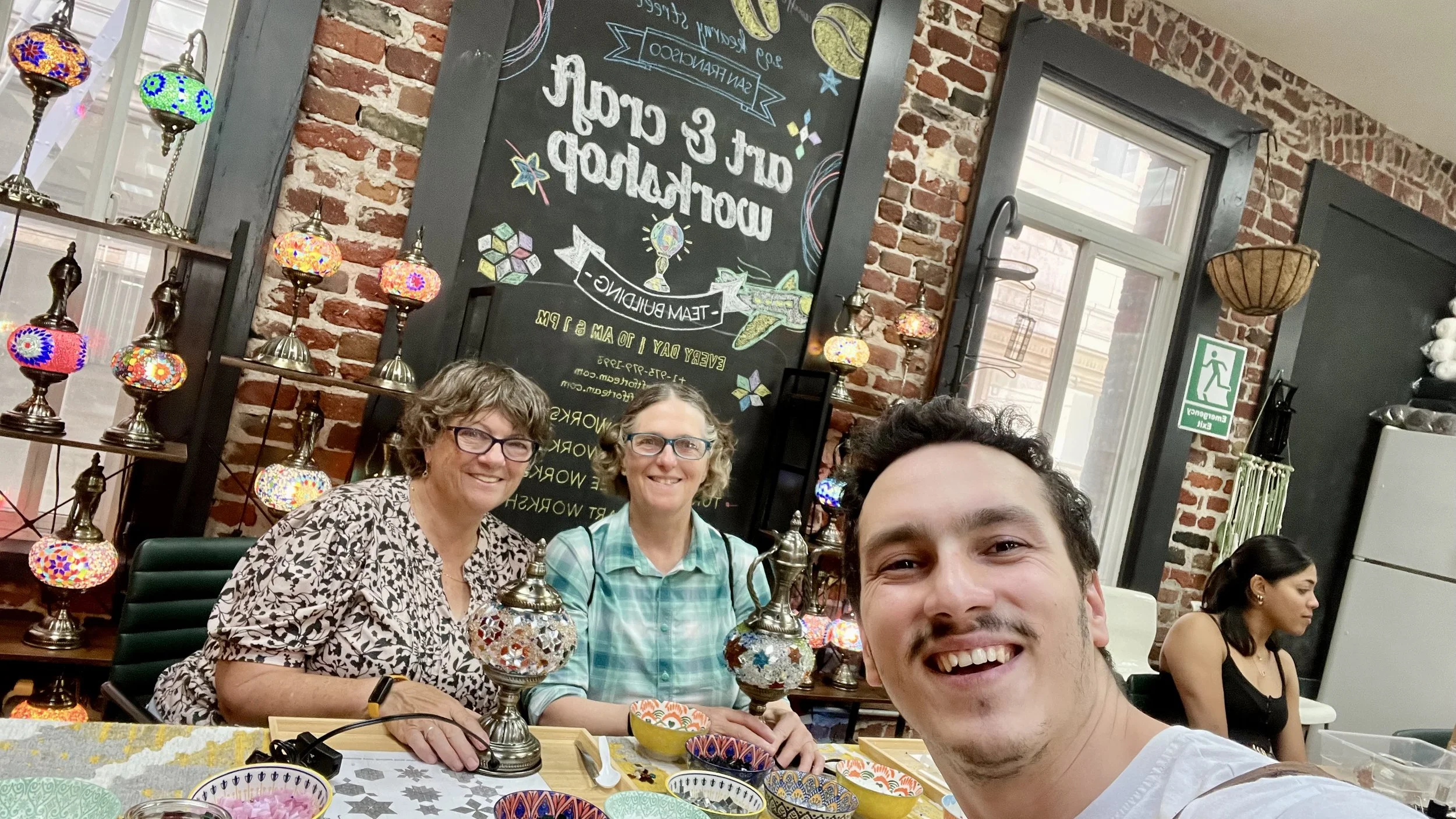 Selfie of three people sitting at a table with colorful lamps and decorative bowls, inside a brick-walled room with a chalkboard sign behind them advertising a workshop or event.