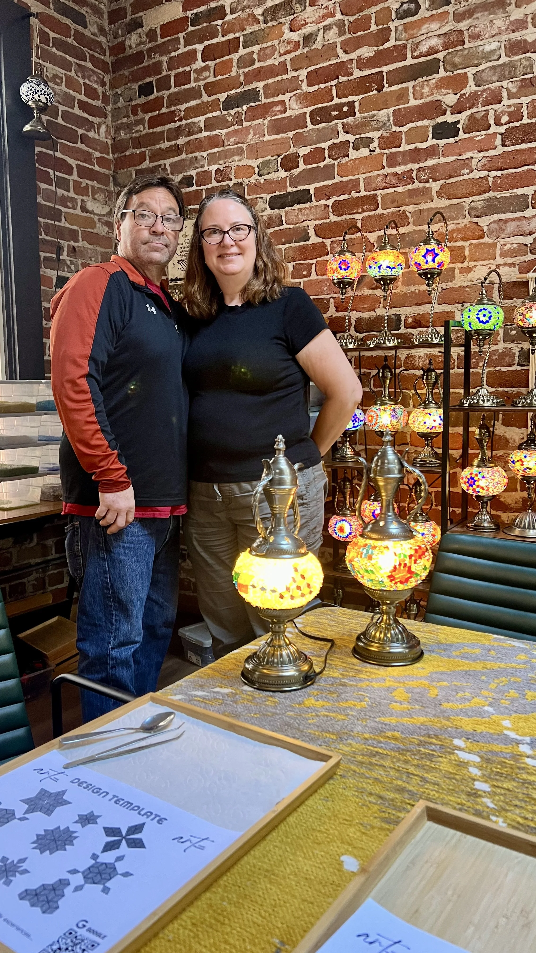 A man and woman standing together in a room with a brick wall, surrounded by colorful mosaic glass lamps on shelves and on a table.