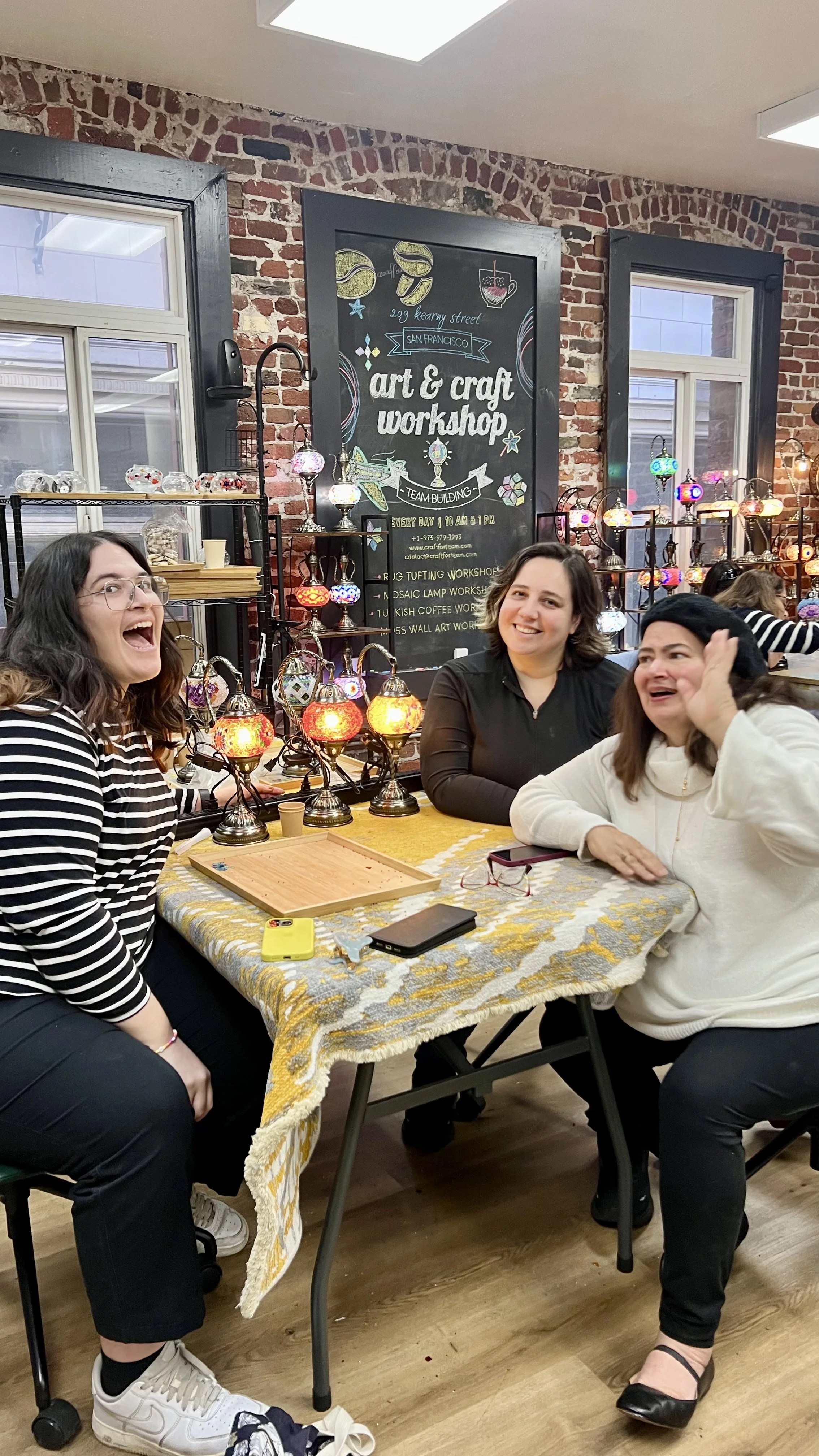 Three women sitting at a table in a craft shop with colorful mosaic lamps behind them, smiling and laughing.