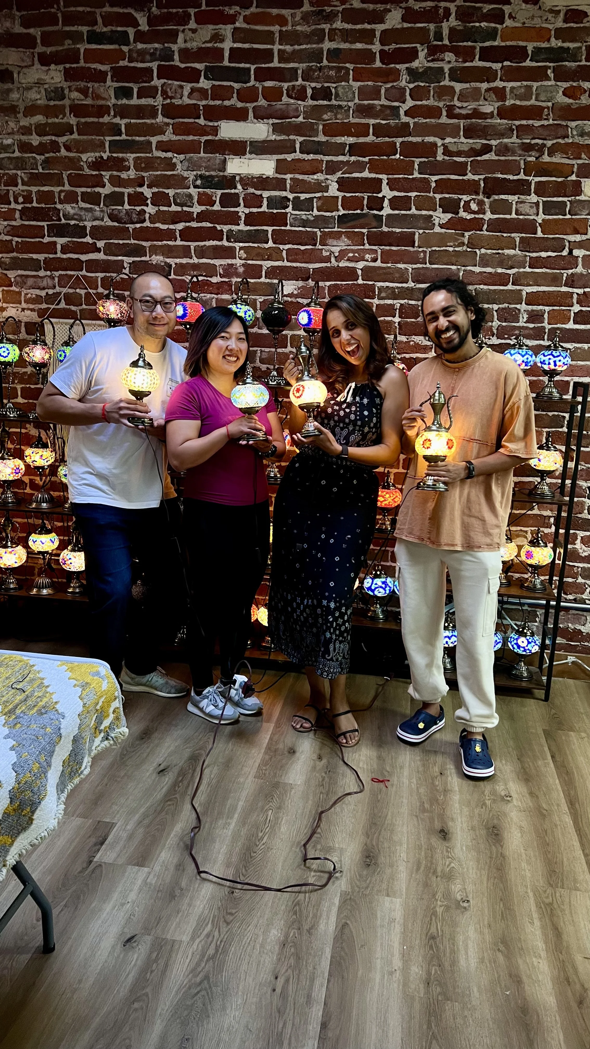 Four people standing in front of a brick wall with colorful Moroccan lamps, holding lamps and smiling.
