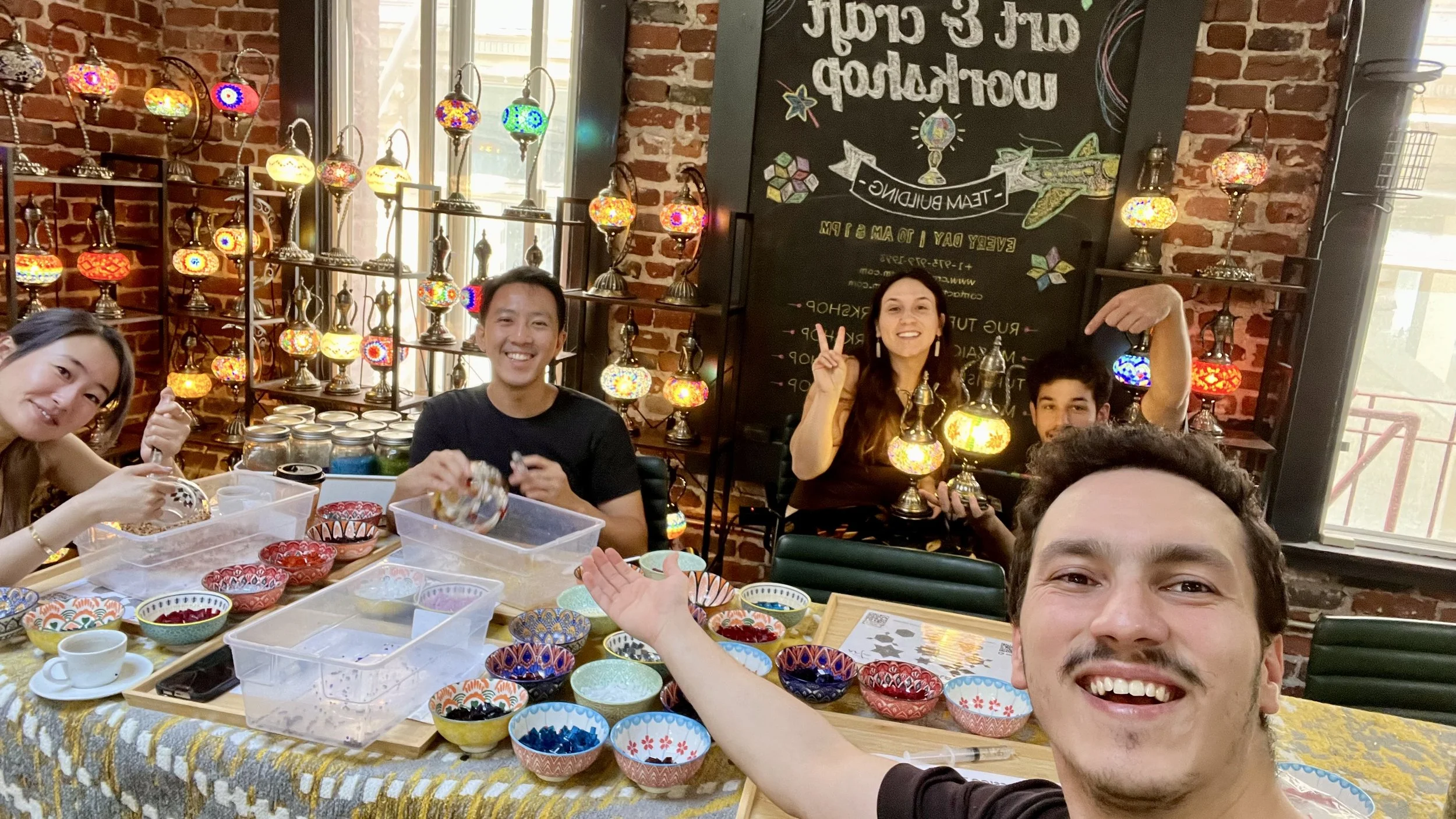 Group of five smiling people sitting around a table with colorful decorated bowls and lamps in a cozy brick-walled shop, celebrating a craft activity.