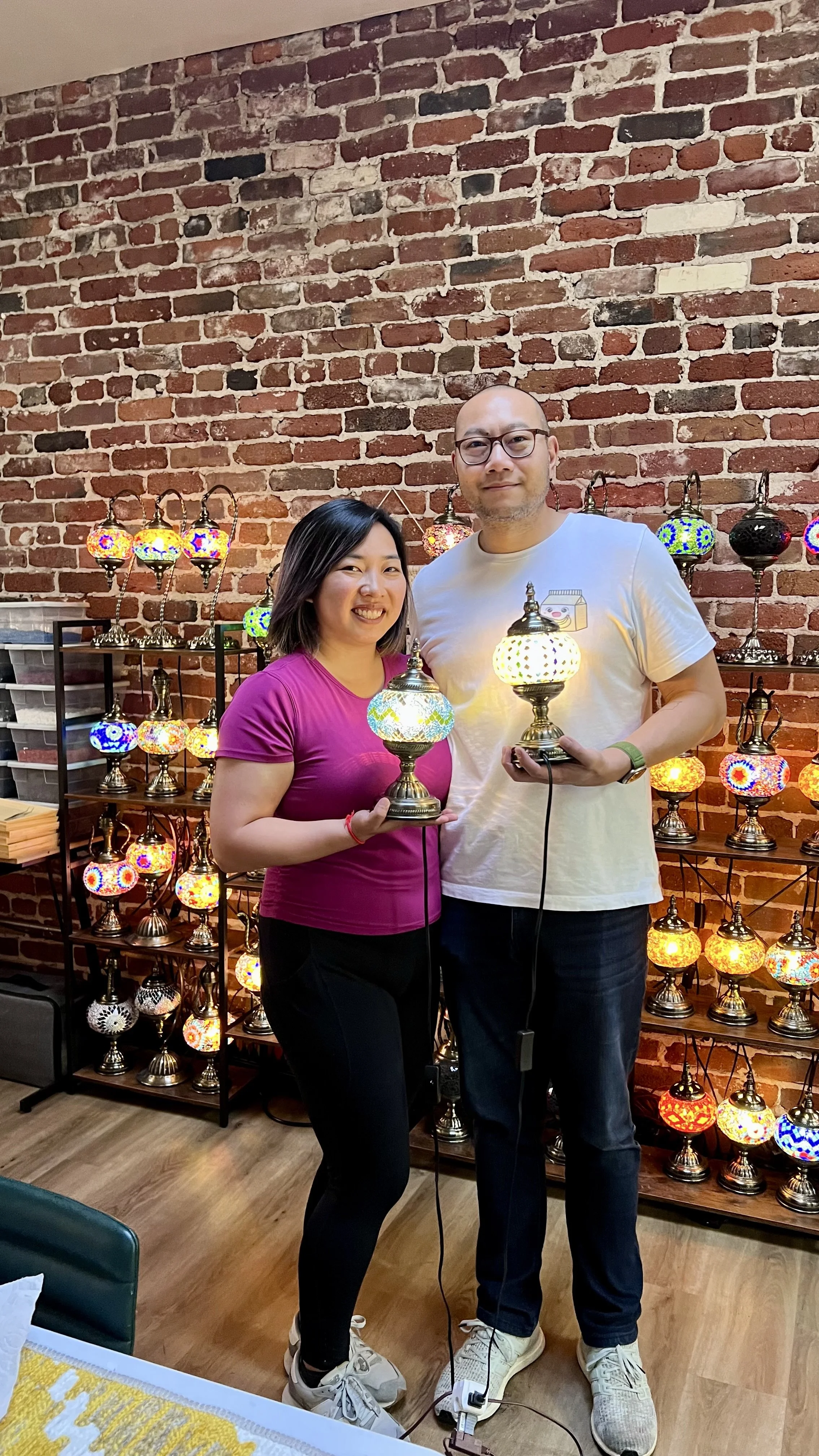 A smiling woman and man holding colorful Moroccan-style lamps in front of a brick wall with a display of decorative lamps.