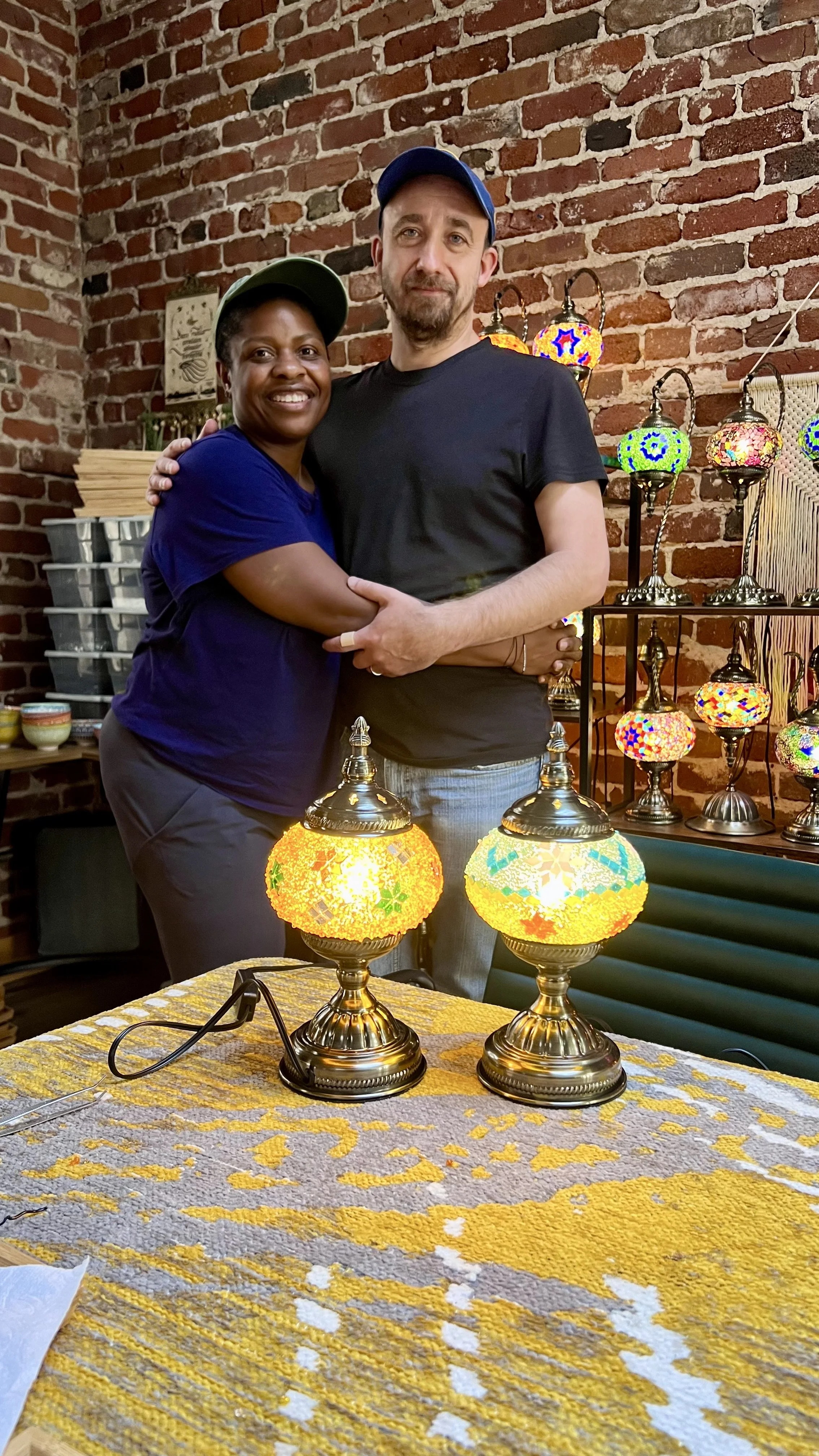 A woman and a man hugging in front of a display of colorful mosaic lamps inside a brick-walled shop.