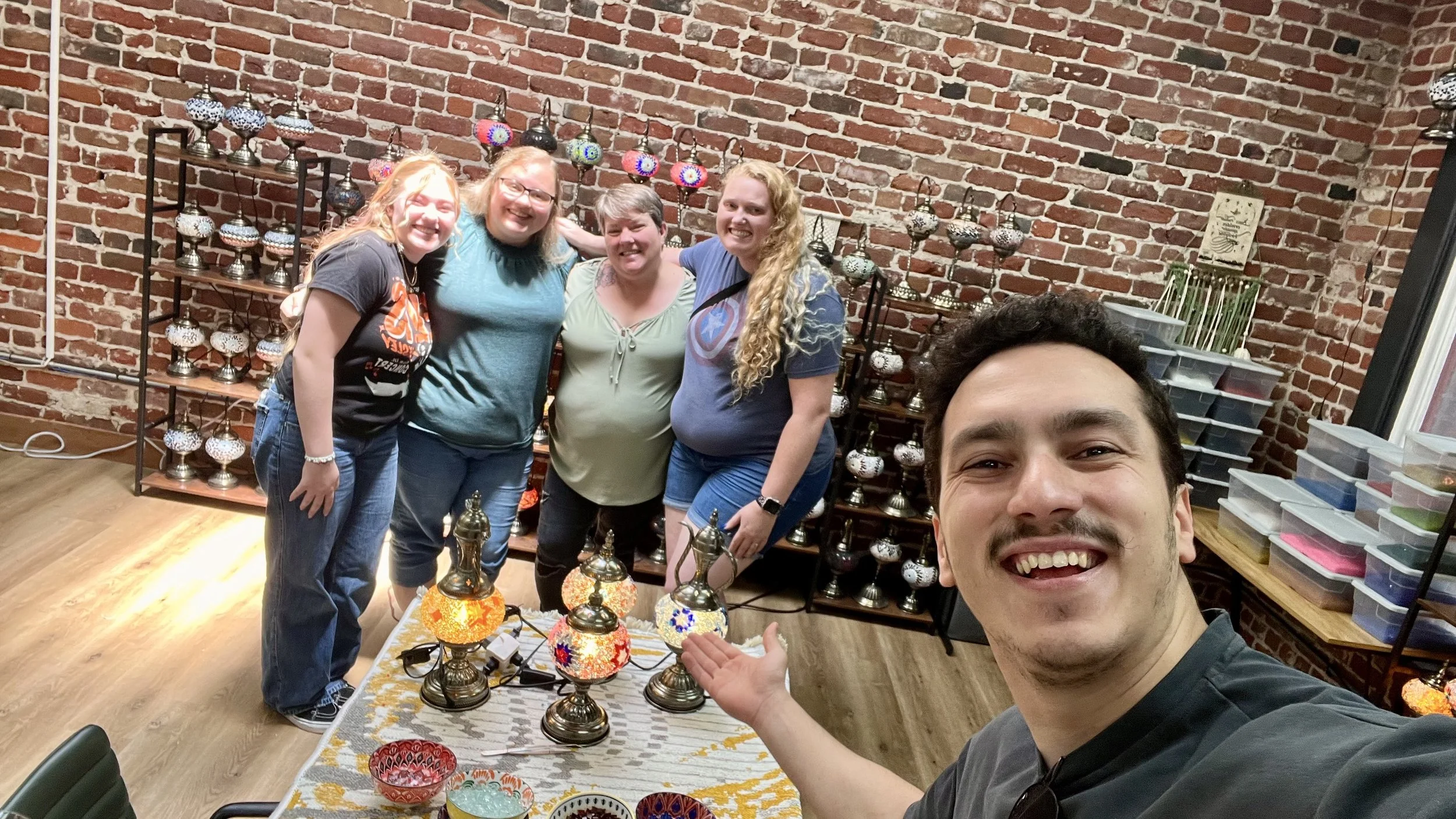 Group of five smiling people taking a selfie in front of a display of colorful Turkish mosaic lamps inside a brick-walled room.