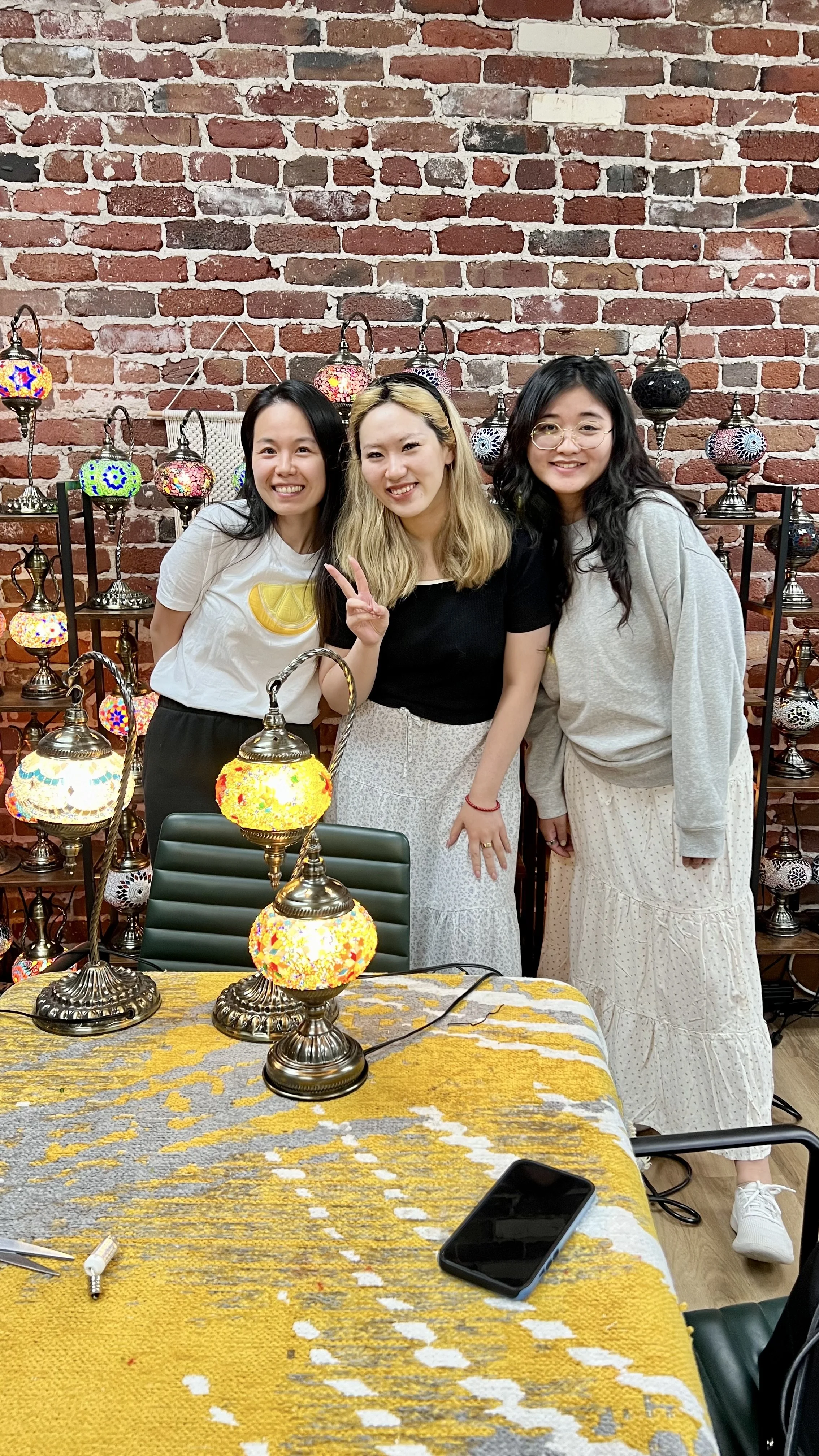 Three women standing behind an ornate table with colorful mosaic lamps, smiling and making peace signs, against a brick wall backdrop.