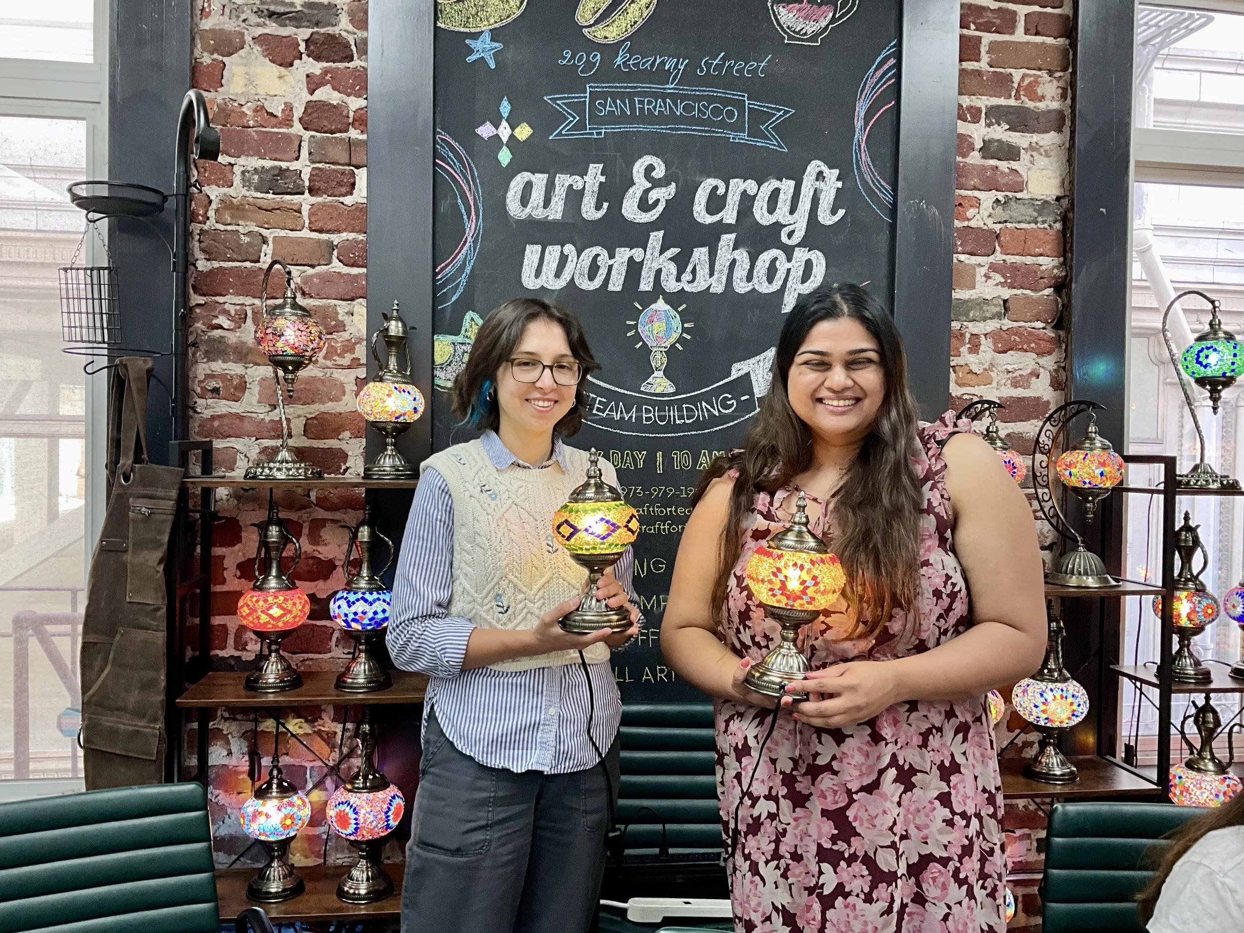 Two women smiling and holding colorful mosaic lamps at an art and craft workshop in front of a brick wall and a chalkboard sign that reads 'art & craft workshop'.