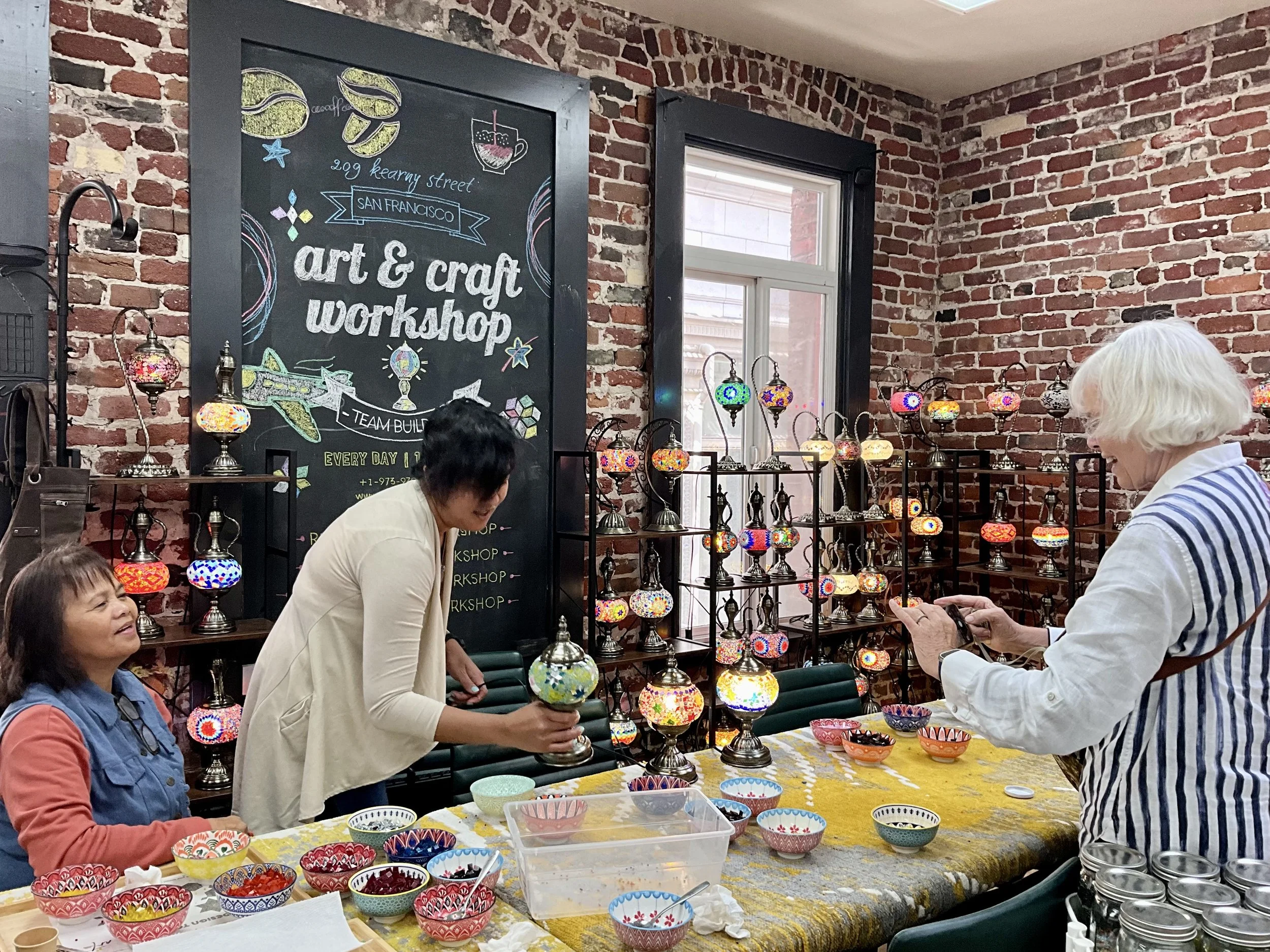 Women in an art and craft workshop creating colorful mosaic lamps, with bowls and craft supplies on a table, brick walls, and shelves displaying finished lamps.