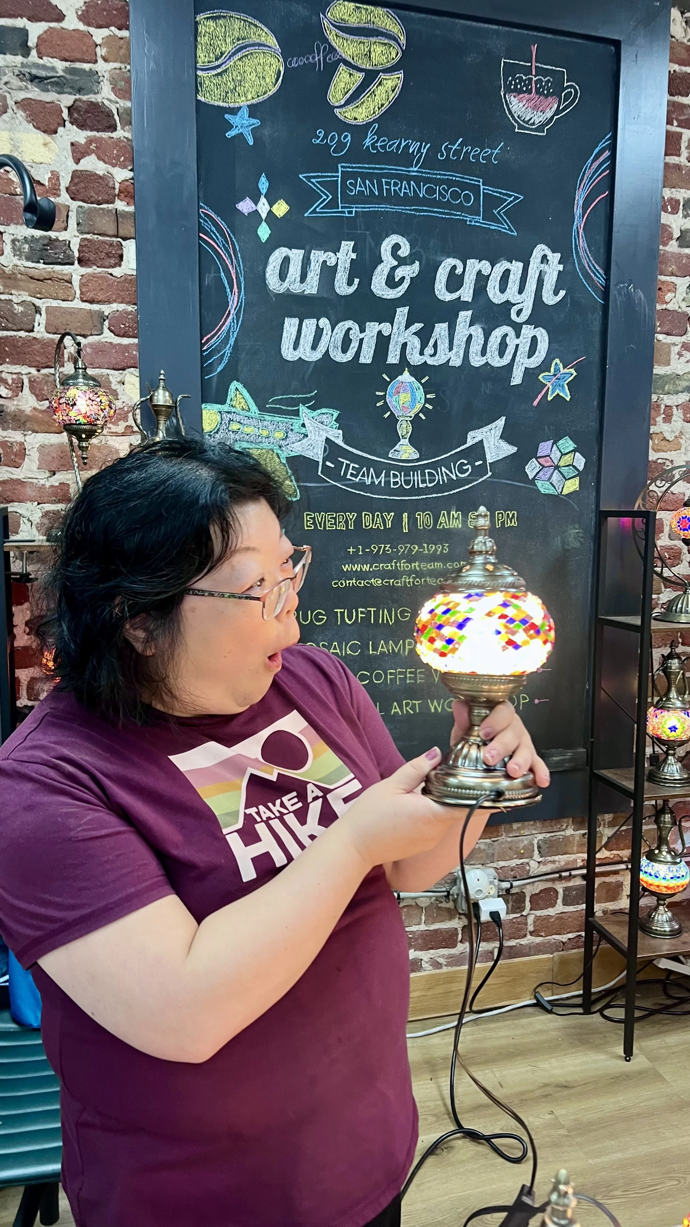 Person holding a colorful mosaic glass lamp in front of a chalkboard advertising an art and craft workshop at 209 Kearny Street in San Francisco, with a brick wall in the background.