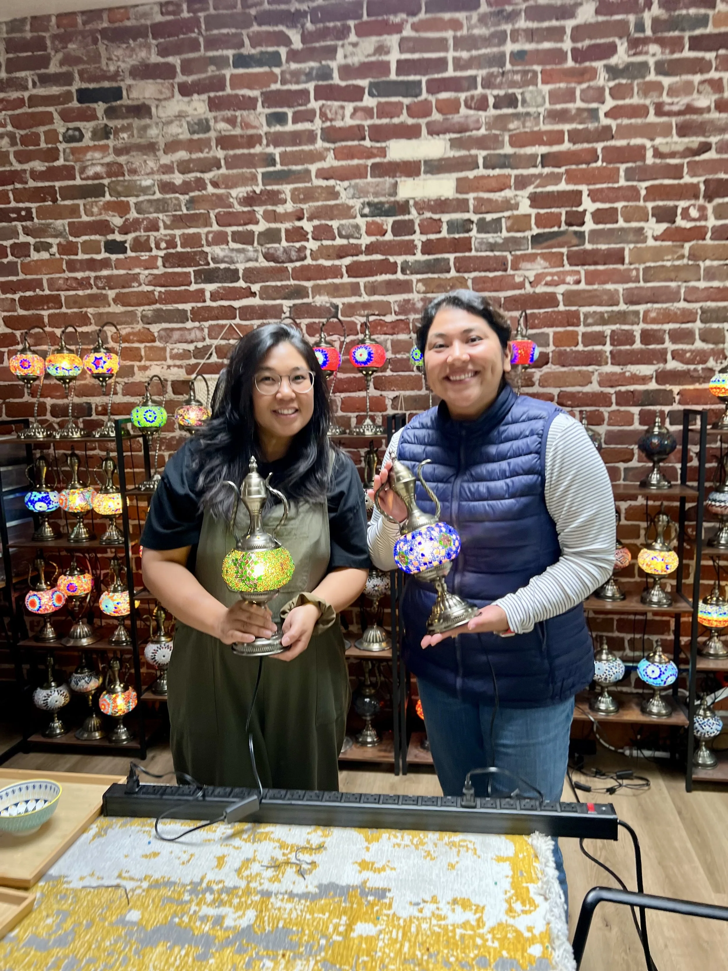 Two smiling women holding colorful mosaic lamps in front of a brick wall with a shelf of similar lamps behind them.