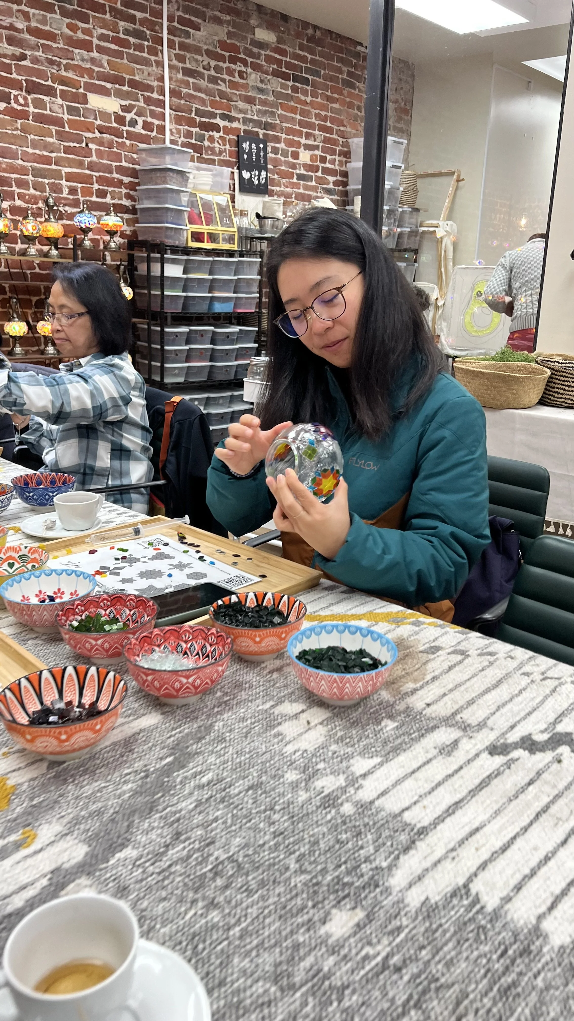 A woman sitting at a table decorating a glass ornament with small colorful tiles; other bowls of mosaic tiles are on the table, along with a teacup and pattern sheets, in a workshop with a brick wall and shelves in the background.