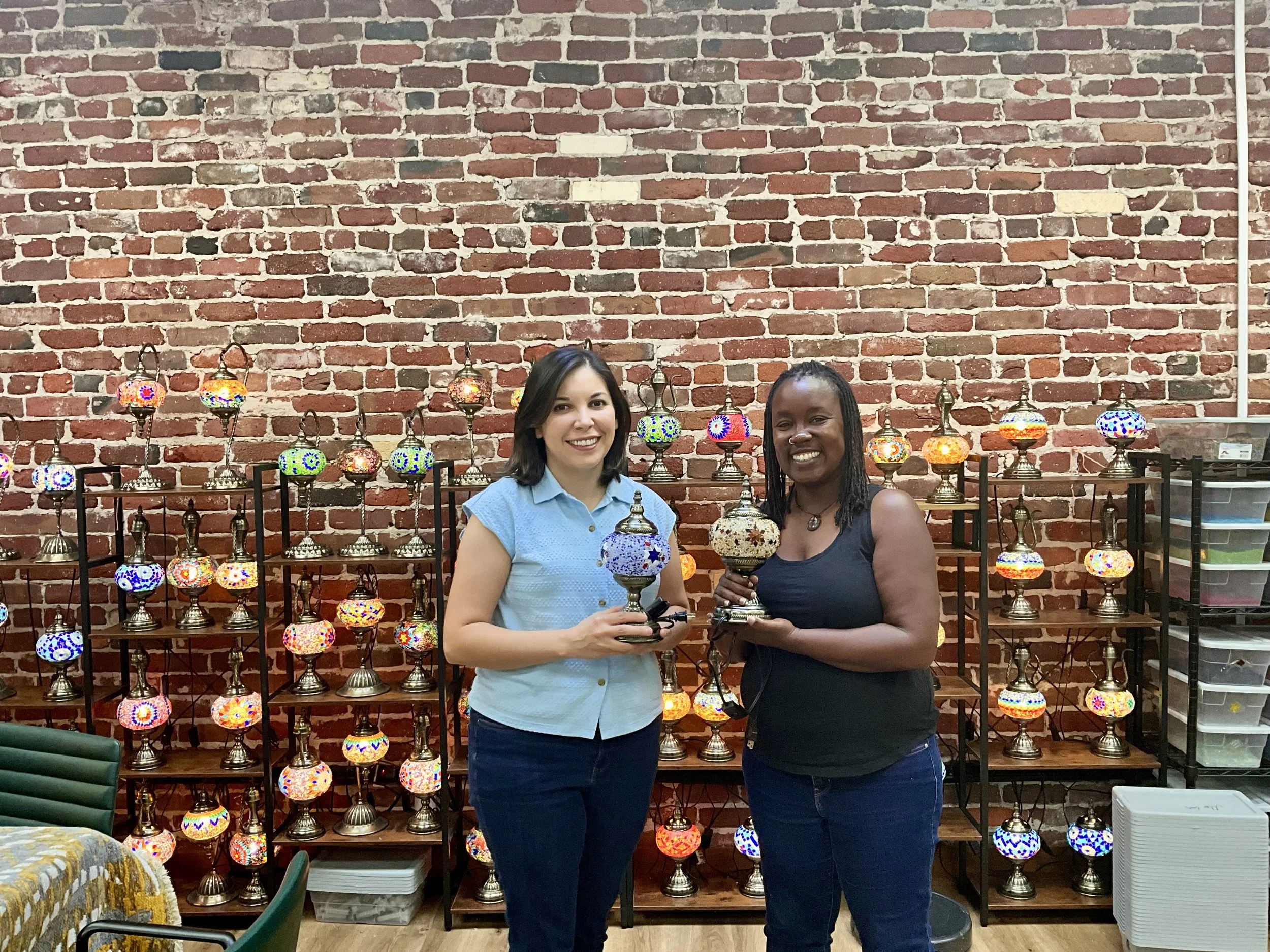 Two women standing in front of a brick wall with colorful mosaic glass lamps on shelves behind them, holding decorative lamps they made.