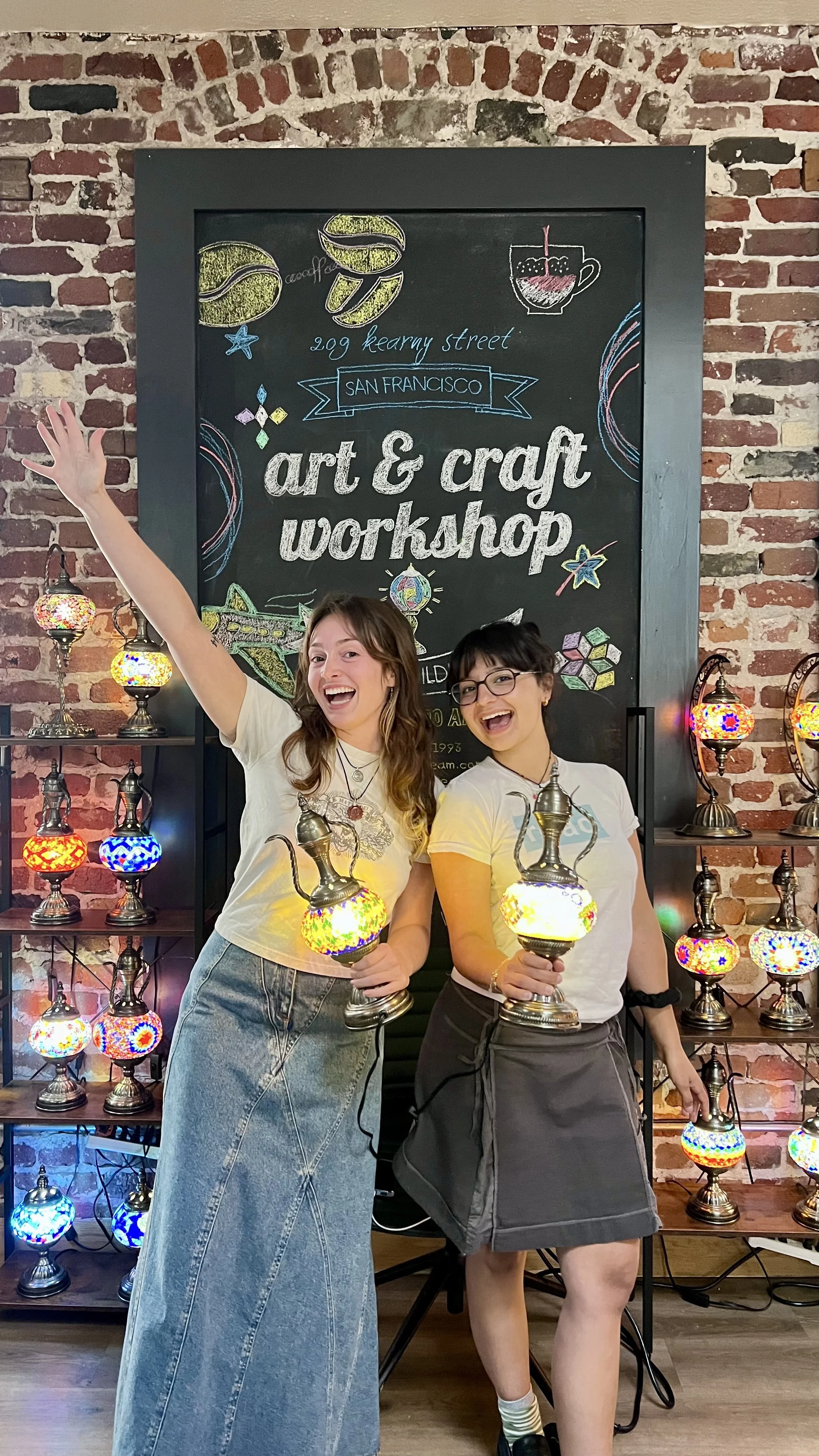 Two women smiling and holding colorful lamps in front of a brick wall and a chalkboard sign that reads 'art & craft workshop' at an indoor craft event.