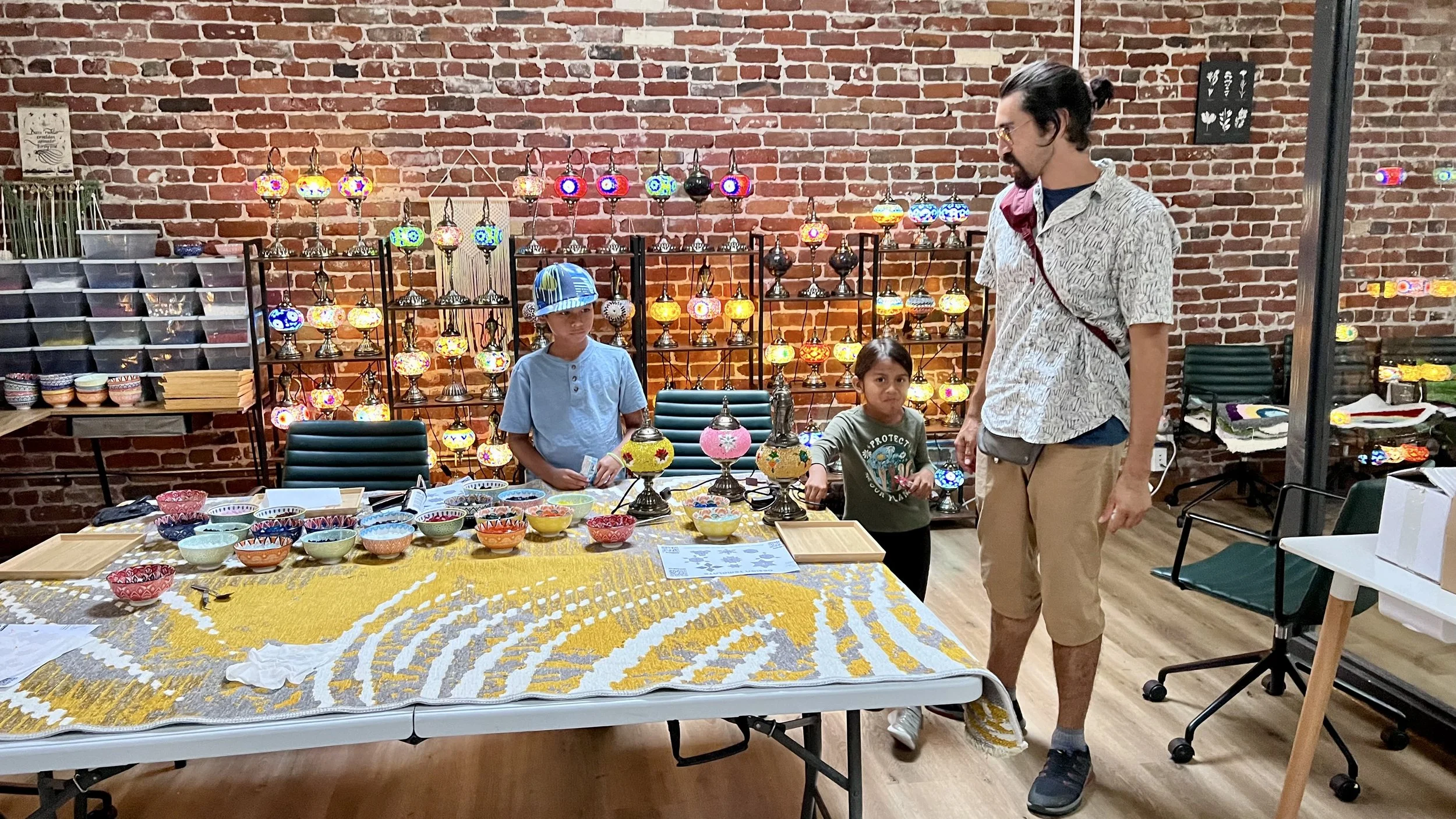 Indoor craft shop with a brick wall background displaying colorful mosaic glass lamps and a table with ceramic bowls. Two children and an adult are looking at the items.