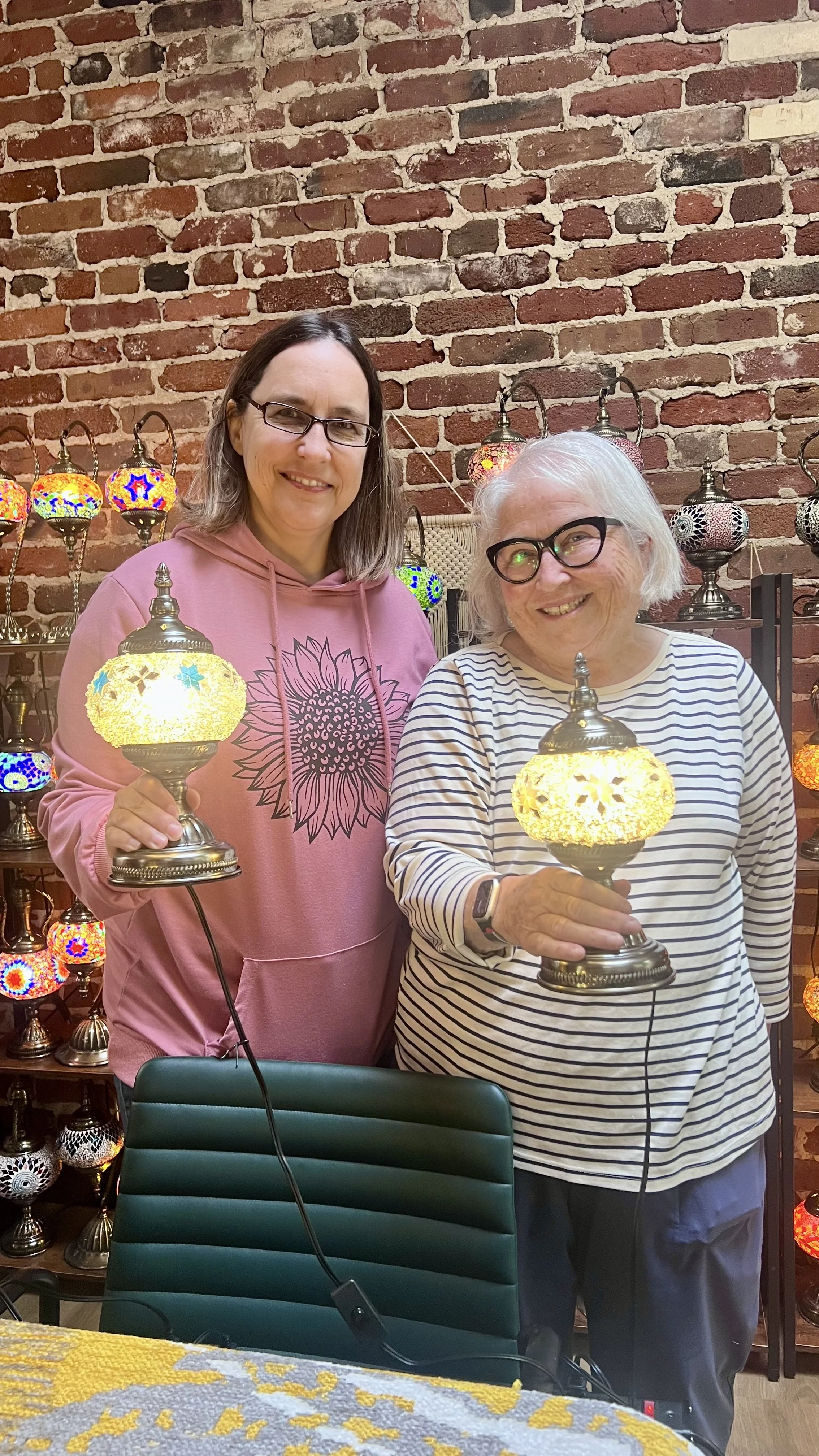 Two women holding decorative table lamps with multicolored glass shades, standing in front of a brick wall display of similar lamps in a shop.