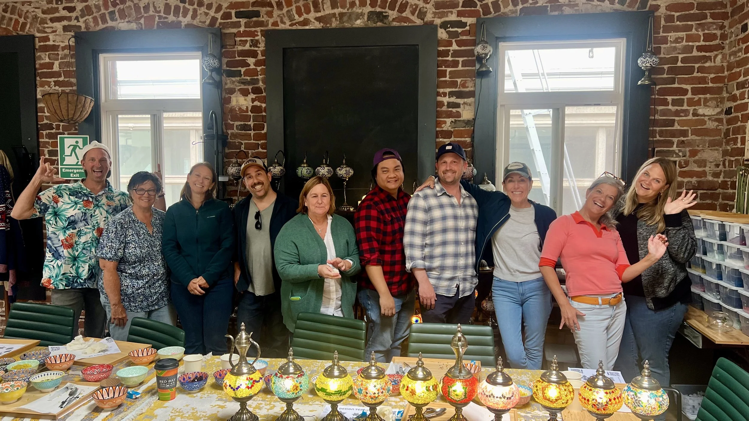 Group of people standing in a room with red brick walls and large windows, smiling and posing for a photo. There are colorful mosaic lamps on the table in front of them.
