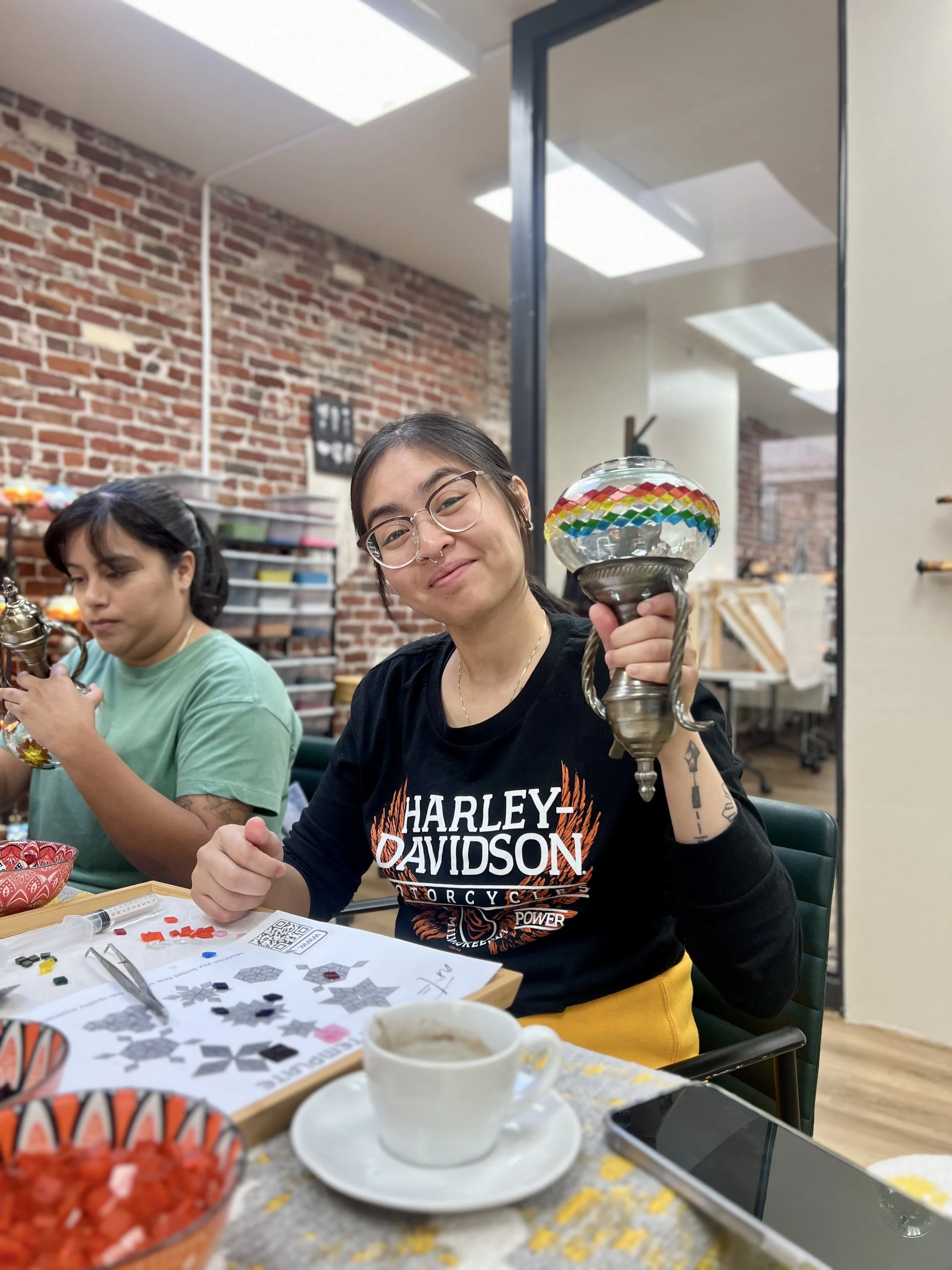 A young woman smiling and holding a colorful glass lamp, seated at a table with crafting supplies, a coffee cup, and a smartphone, with a brick wall and another person in the background.
