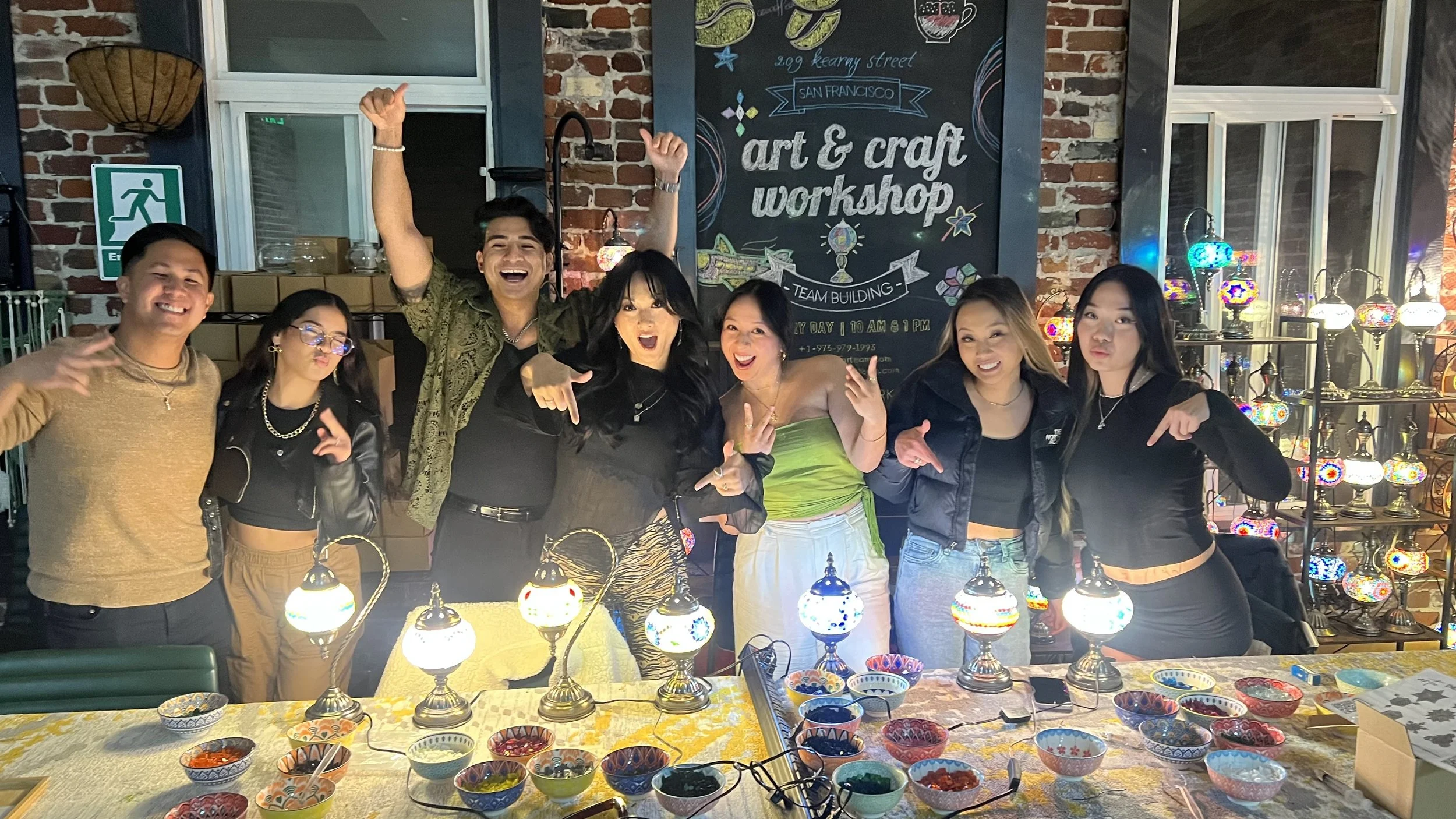 Group of seven people celebrating at an art and mosaic lamp craft workshop, posing and smiling in front of a chalkboard sign.
