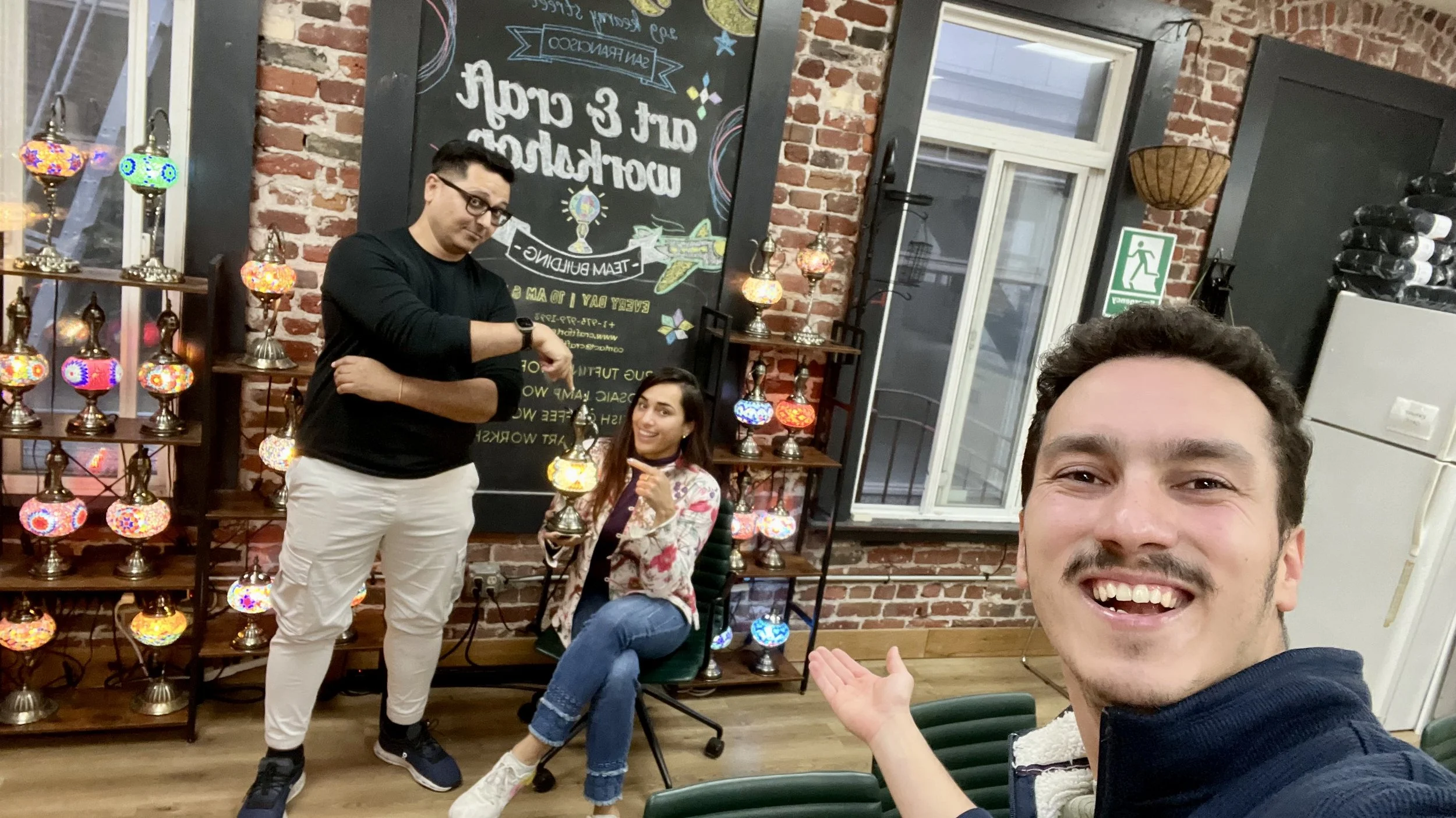 Three individuals in a room with brick walls and a chalkboard, with colorful lamps on shelves; one man takes a selfie with two others, one sitting on a desk and the other standing beside him, smiling and pointing at the seated woman.