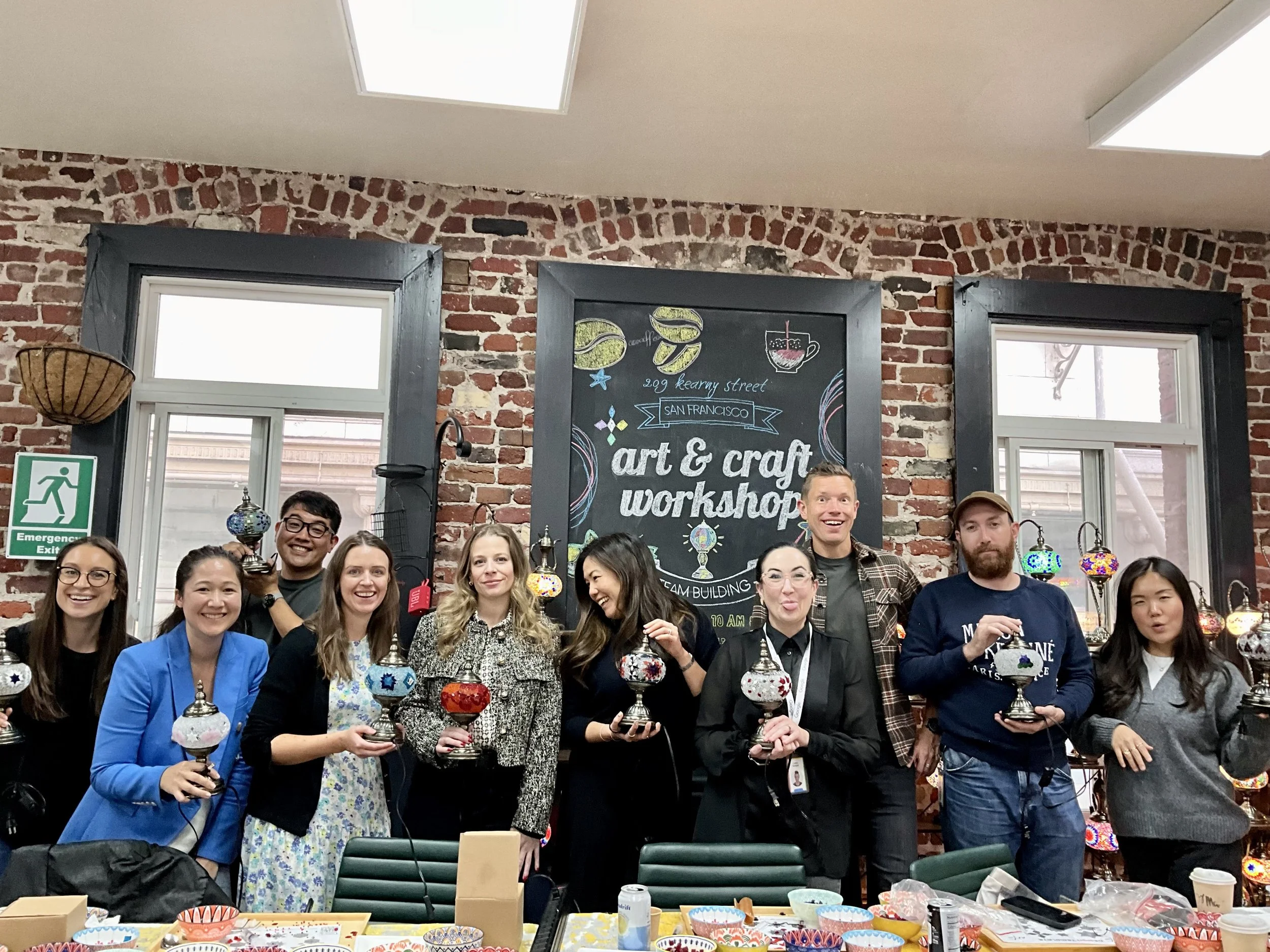 A group of people standing in a craft workshop, holding colorful glass or ceramic ornaments, smiling and posing for a photo inside a brick-walled room with a chalkboard sign that reads 'art & craft workshop', with windows and displayed crafts in the 