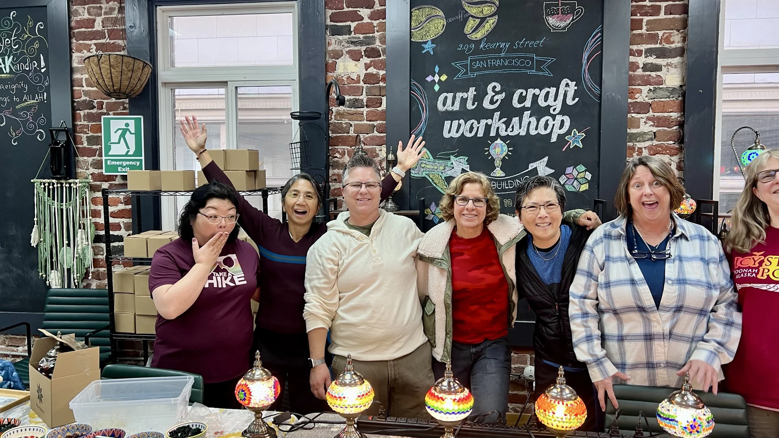 Group of women smiling and celebrating at an art and craft workshop, standing behind a table with colorful lamps and craft supplies, in a brick-walled studio with a chalkboard sign that reads 'art & craft workshop'.