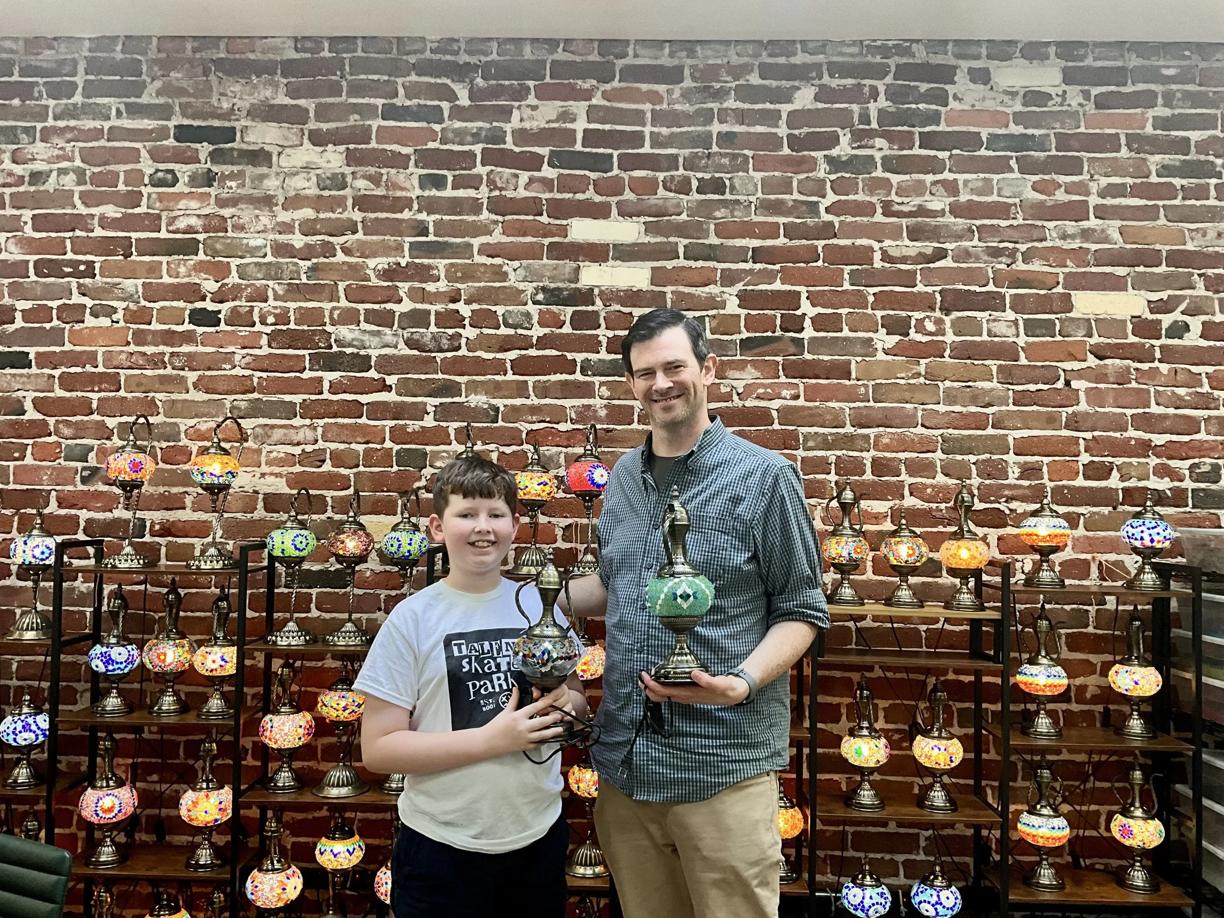 Two males, a boy and a man, standing in front of a brick wall and a display of colorful mosaic lamps, holding trophies and smiling.